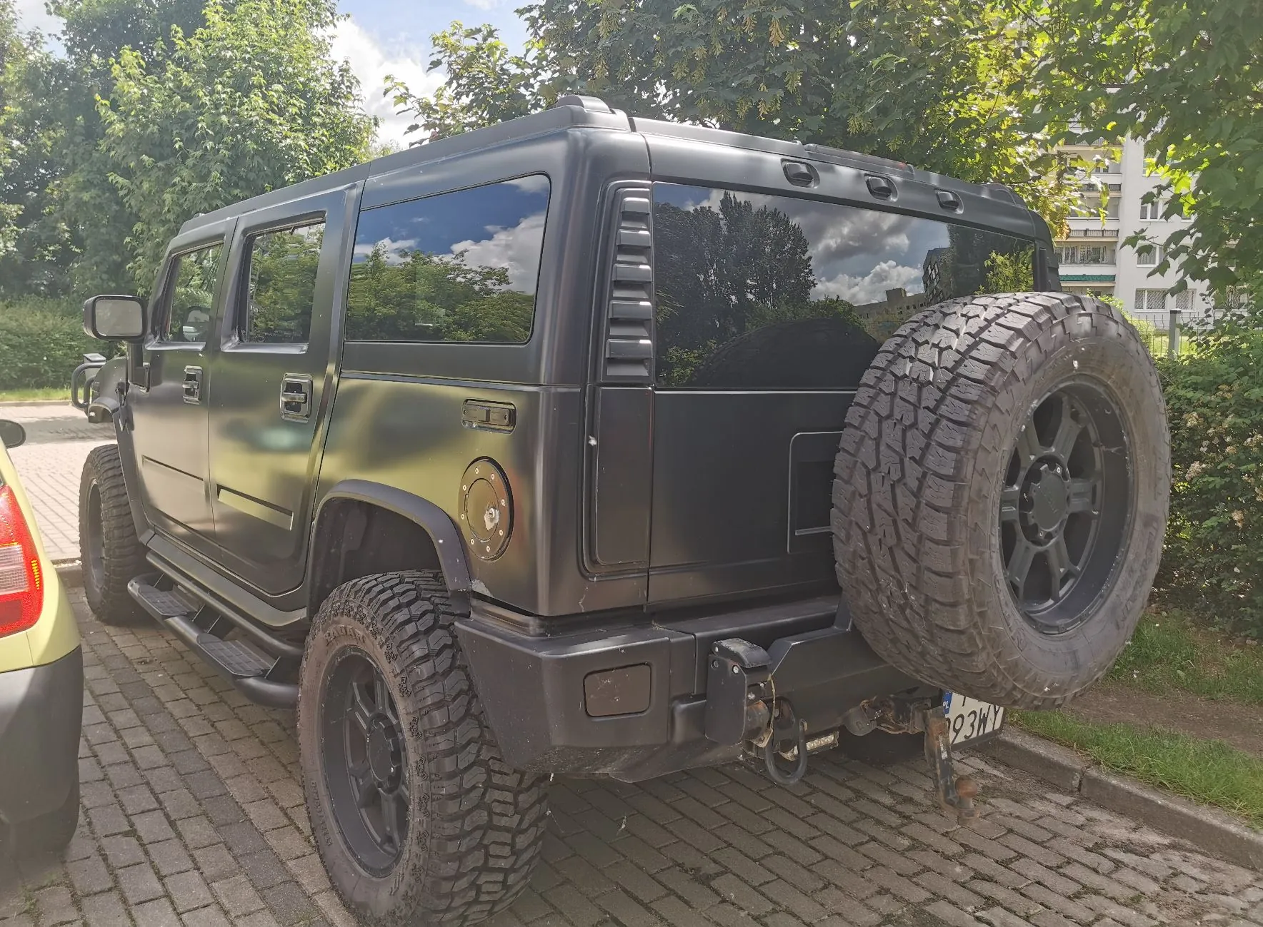 Czarny, terenowy samochód typu Hummer zaparkowany na kostce brukowej. Pojazd ma duże, terenowe opony z czarnymi felgami, jedną z nich zamontowano na tylnej klapie. Samochód posiada przyciemniane szyby i jest ustawiony bokiem do obiektywu, ukazując tylną i lewą stronę nadwozia. W tle widoczne są zielone drzewa oraz fragment białego budynku mieszkalnego.