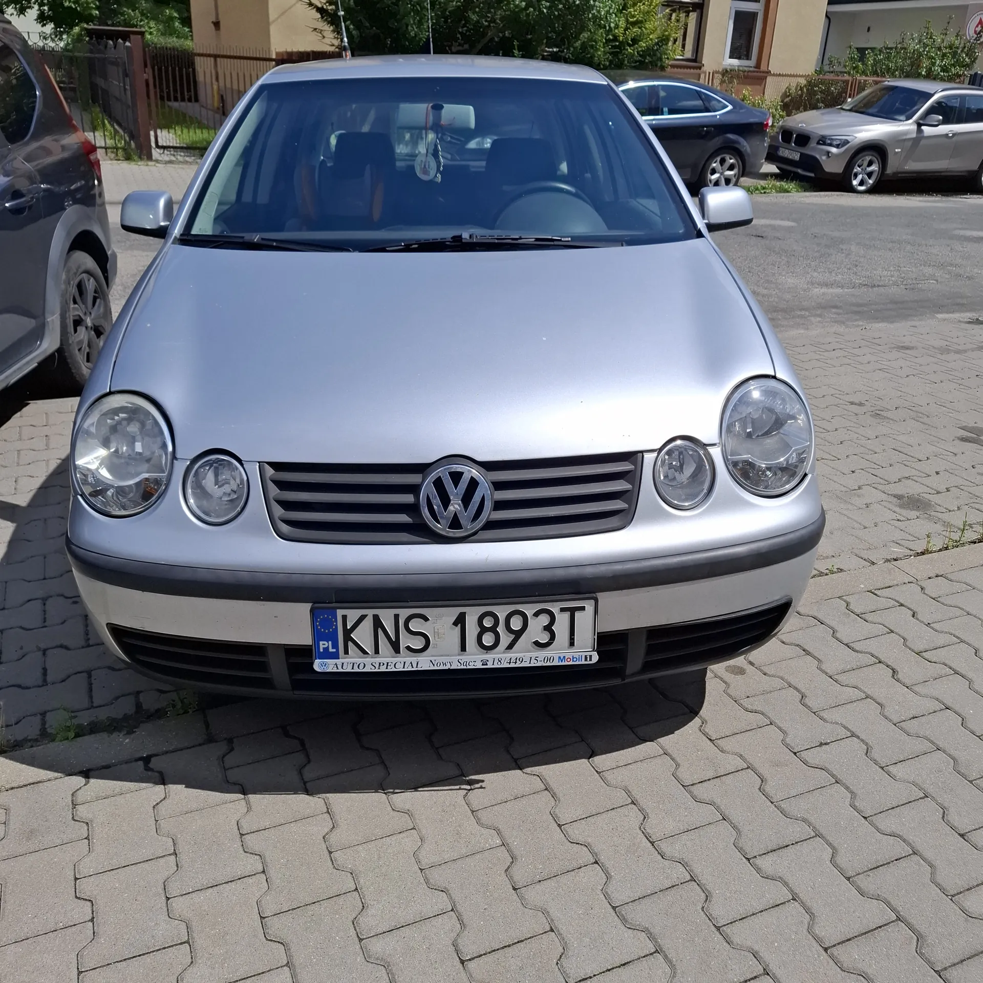 Przód srebrnego samochodu marki Volkswagen z tablicą rejestracyjną KNS 1893T. Widoczne są reflektory, logo marki oraz inne zaparkowane pojazdy na tle budynków i drzew. Tablica rejestracyjna zawiera dodatkowe napisy: PL, AUTO SPECIAL, numer telefonu 18 449-15-00 oraz nazwę firmy.