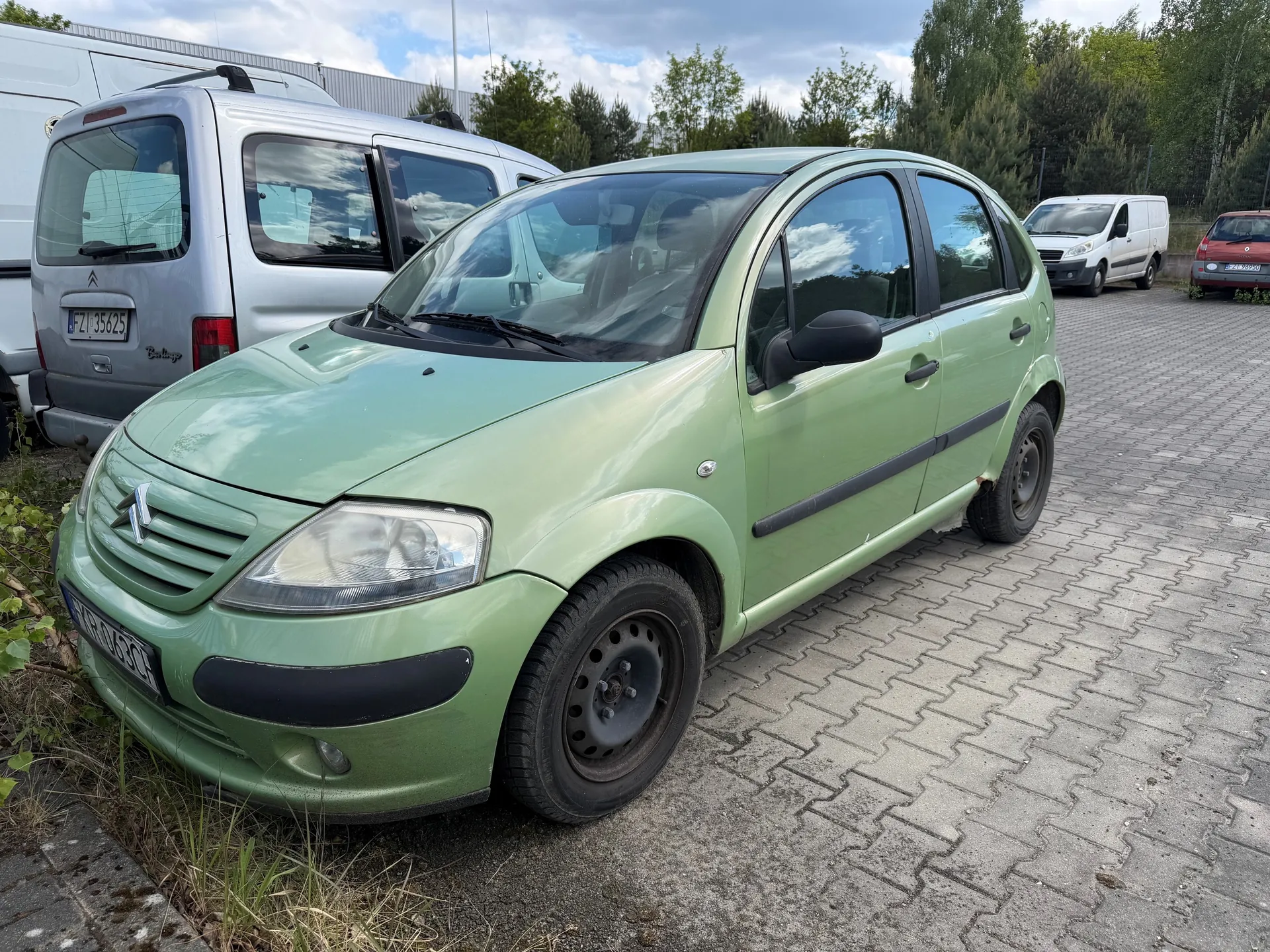 Zielony samochód Citroën z tablicą rejestracyjną WRO 03C zaparkowany na brukowanym placu. Za nim srebrny furgon Citroën z tablicą FZ 35625. W tle białe furgony, czerwony samochód, drzewa i budynek. Niebo częściowo zachmurzone.