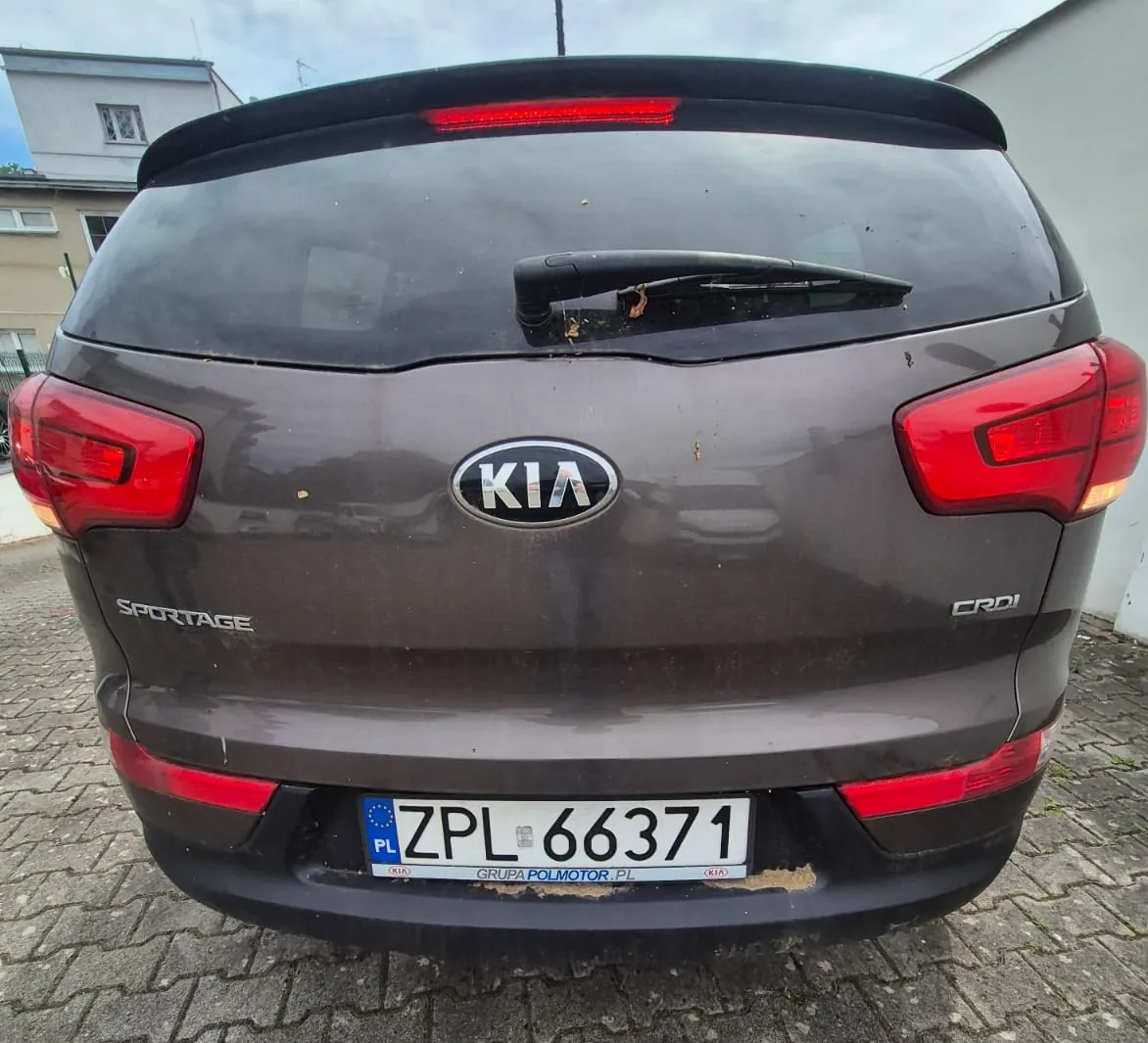 Tylny widok brązowego samochodu marki Kia, model Sportage. Na klapie bagażnika znajduje się logo KIA, napis SPORTAGE po lewej stronie oraz oznaczenie CRDi po prawej. Samochód ma polską tablicę rejestracyjną o numerze ZPL 66371, z ramką Grupa Polmotor.pl. Pojazd jest lekko zabrudzony.