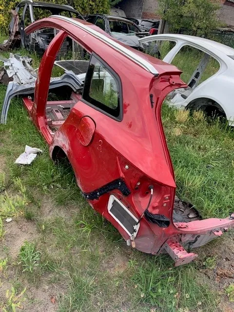 Fragment czerwonej karoserii samochodu, obejmujący tylną część nadwozia z drzwiami i dachem, leżący na trawie na złomowisku. W tle widoczne są inne części samochodowe, w tym białe nadwozie auta oraz ciemne wraki.