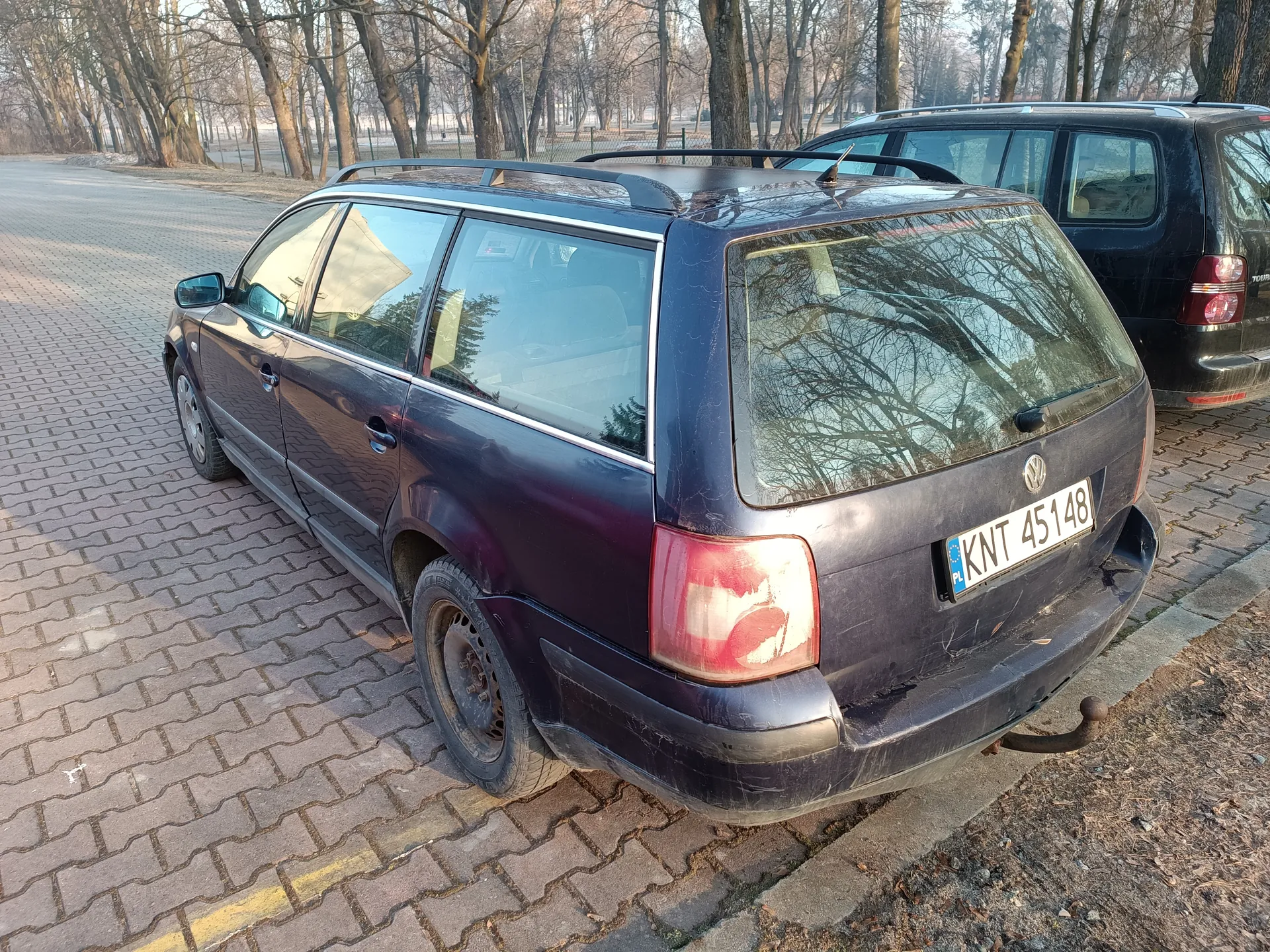 Ciemnofioletowy samochód osobowy typu kombi marki Volkswagen zaparkowany na brukowanej powierzchni. Widoczny tył pojazdu z tablicą rejestracyjną KNT 45148. W tle czarny samochód oraz drzewa bez liści. Parkowanie na obszarze z brukowaną drogą i żółtą linią wyznaczającą pas.