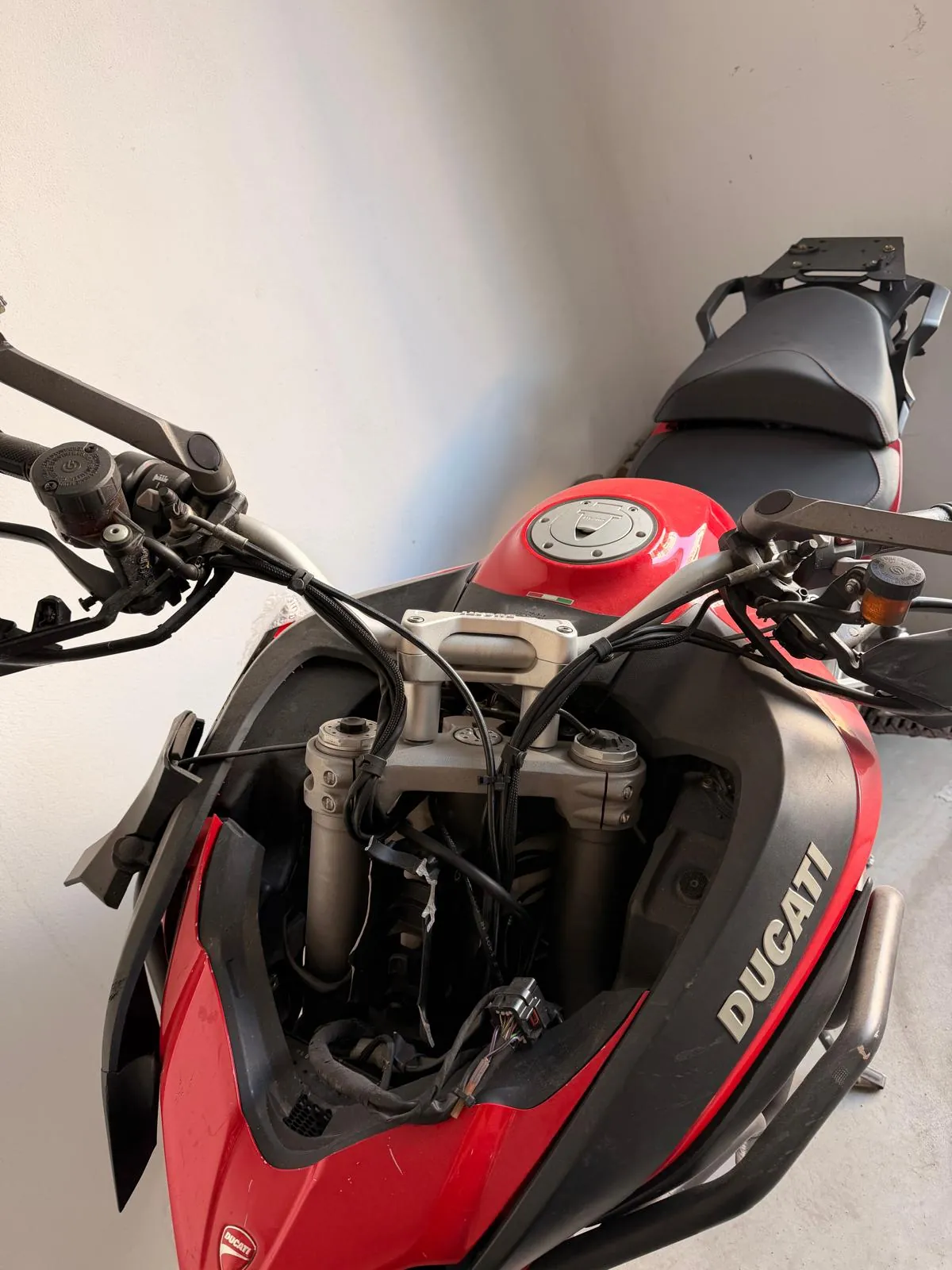 Zbliżenie na przednią część czerwono-czarnego motocykla marki Ducati. Widoczna jest kierownica z manetkami, zbiornik paliwa z wlewem, przedni zawieszenie oraz fragment siedzenia. Na boku owiewki znajduje się biały napis 'DUCATI'.