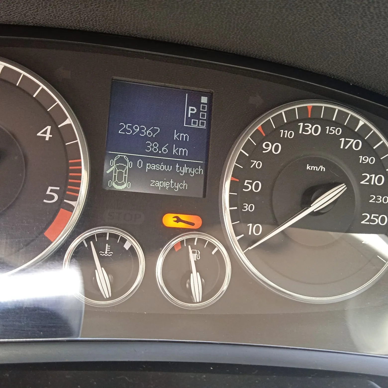 Zbliżenie na deskę rozdzielczą samochodu. W centrum znajduje się cyfrowy wyświetlacz z tekstem: '259367 km', '38.6 km' oraz '0 pasów tylnych zapiętych' obok grafiki samochodu. Poniżej wyświetlacza widnieje pomarańczowa kontrolka w kształcie klucza nasadowego. Po lewej stronie znajduje się obrotomierz z widocznymi cyframi 4 i 5, a po prawej prędkościomierz z podziałką do 250 km/h. Na dole widoczne są dwa analogowe wskaźniki: temperatury cieczy chłodzącej oraz poziomu paliwa.