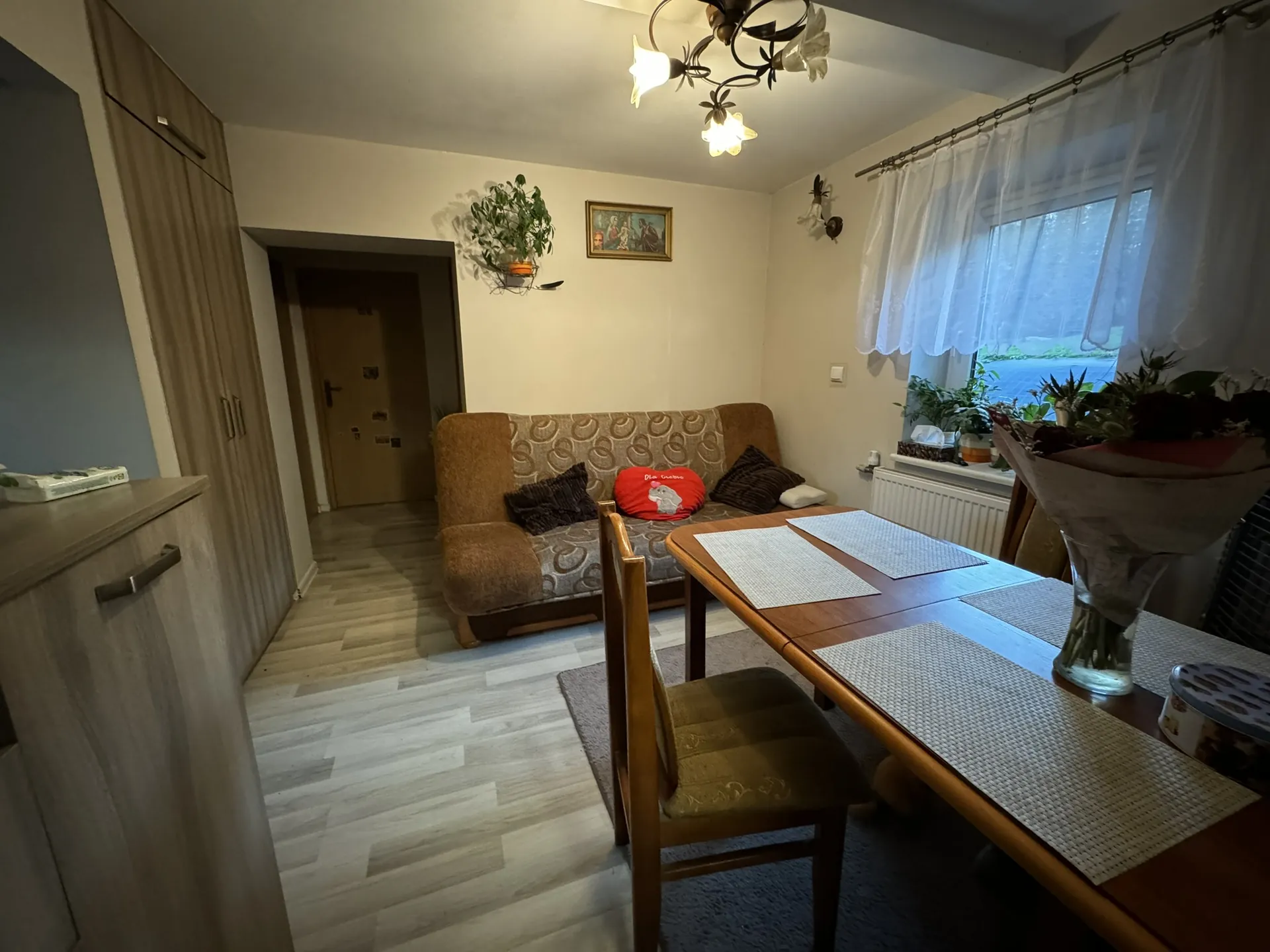 Salon z brązową sofą o wzorzystej tapicerce, na której leżą dwie ciemne poduszki i czerwona poduszka z napisem „Dla Ciebie”. Przed sofą stół obiadowy z czterema podkładkami, szklanym wazonem z bukietem kwiatów i pojemnikiem. Po prawej stronie okno z białymi koronkowymi zasłonami, na parapecie rosną rośliny. Na ścianie nad sofą wiszą obraz w ramie i wisząca doniczka z rośliną. W lewej części pokoju szafy oraz komoda z przedmiotami. Podłoga wyłożona jasnymi płytkami imitującymi drewno. Oświetlenie zapewnia żyrandol z zapalonymi lampami.