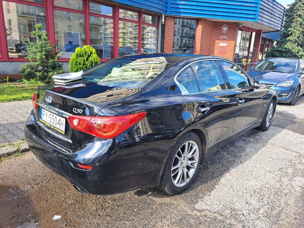 Czarny samochód marki Infiniti model Q50 zaparkowany na podjeździe przed budynkiem. Widoczny jest tył i prawy bok auta. Samochód ma polską tablicę rejestracyjną o numerze PY 5504H. Na tylnej półce wewnątrz auta znajdują się jasne przedmioty. W tle widać nowoczesny budynek z dużymi przeszklonymi witrynami oraz niebieskim elementem elewacji na górze.
