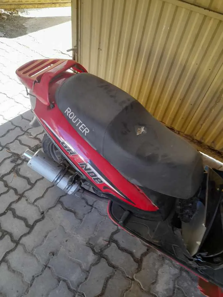 Czerwono-czarny skuter z czarnym siedzeniem, na którym widnieje biały napis ROUTER. Na boku skutera znajduje się napis GRAND. Skuter stoi na brukowanej powierzchni obok pomarańczowej, pionowo blachowanej ściany. W tle widoczna jest jasna, słoneczna przestrzeń z częścią nawierzchni i roślinnością.