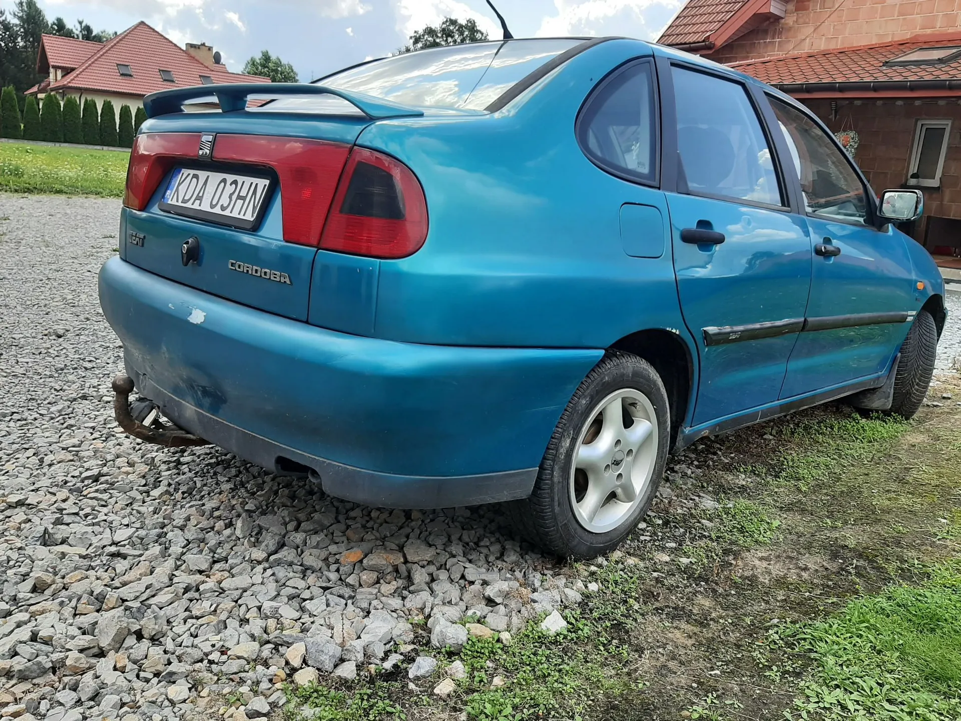 Niebieski samochód Seat Cordoba zaparkowany na żwirowym podjeździe. Widoczny jest tył i prawy bok pojazdu. Samochód posiada polską tablicę rejestracyjną o numerze KDA 03HN oraz napis CORDOBA na klapie bagażnika. W tle widać domy z czerwonymi dachami i zieloną roślinność.