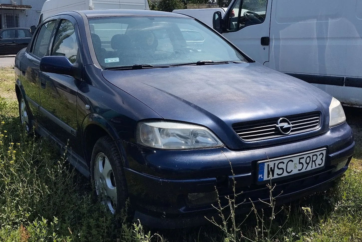 Granatowy samochód osobowy marki Opel zaparkowany na trawie. Widoczna jest przednia część pojazdu z tablicą rejestracyjną o numerze WSC 59R3. W tle znajduje się biały samochód dostawczy.