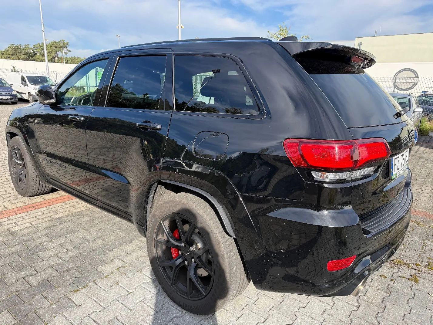 Czarny samochód typu SUV zaparkowany na kostce brukowej. Zdjęcie przedstawia tył i prawy bok pojazdu. Samochód ma czarne felgi z czerwonymi zaciskami hamulcowymi. W tle widoczne są inne samochody, biały budynek oraz jasne niebo z lekkimi chmurami.