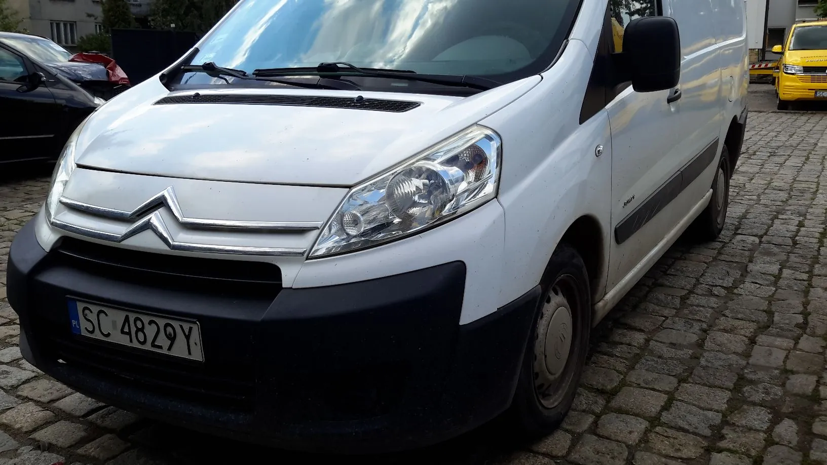 Biały samochód dostawczy marki Citroën zaparkowany na kostce brukowej. Widoczny jest przód pojazdu z tablicą rejestracyjną o treści SC 4829Y. W tle widać fragmenty innych samochodów oraz budynek.