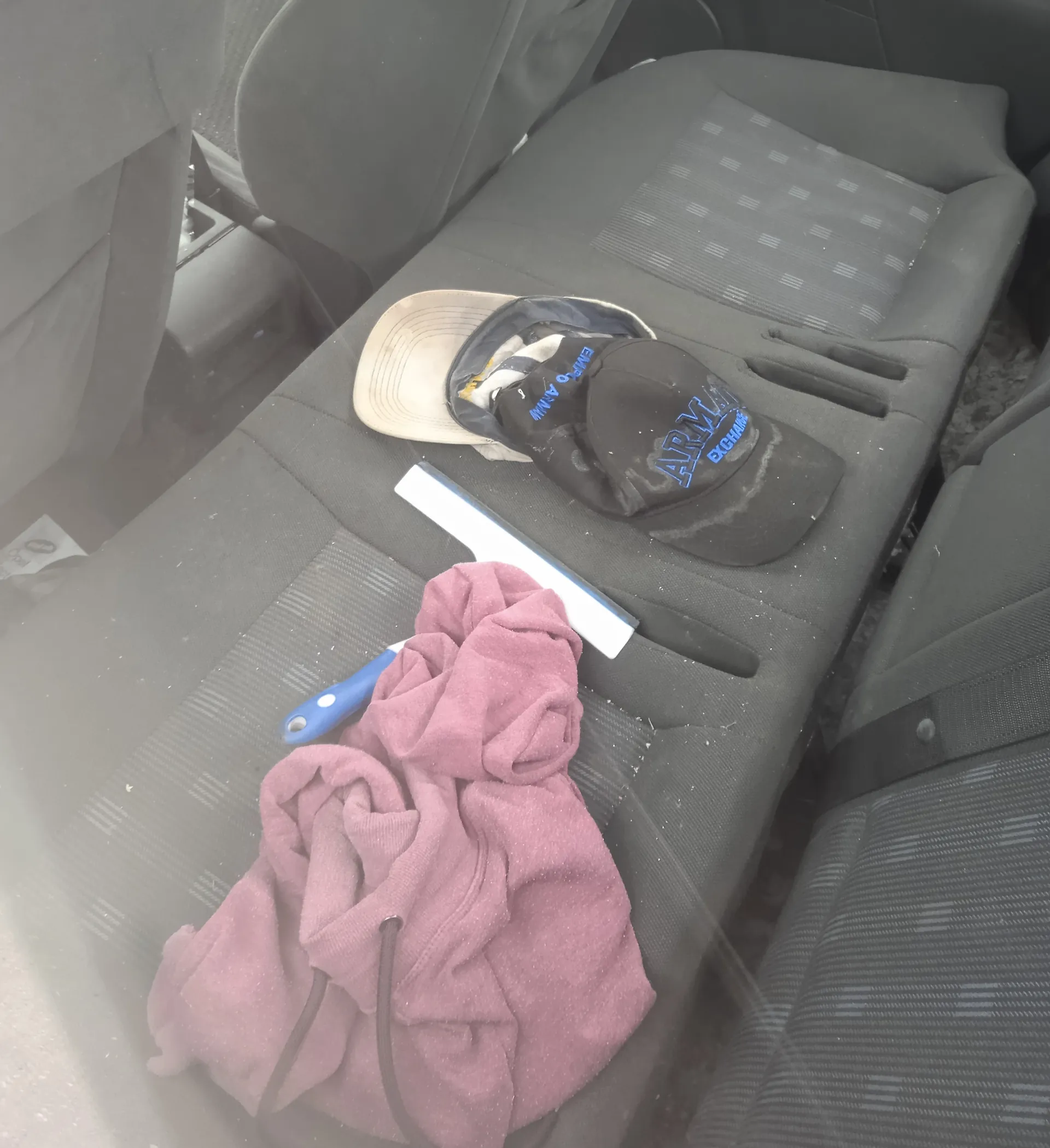 Wnętrze samochodu z widocznym tylnym siedzeniem w kolorze szarym. Na siedzeniu leży różowa bluza z kapturem, czarna czapka z niebieskim napisem "ARMY EXCHANGE", beżowa czapka, biały przedmiot oraz niebiesko-biały przedmiot przypominający skrobak. Widoczne są również fragmenty pasów bezpieczeństwa i siatek na siedzeniach.