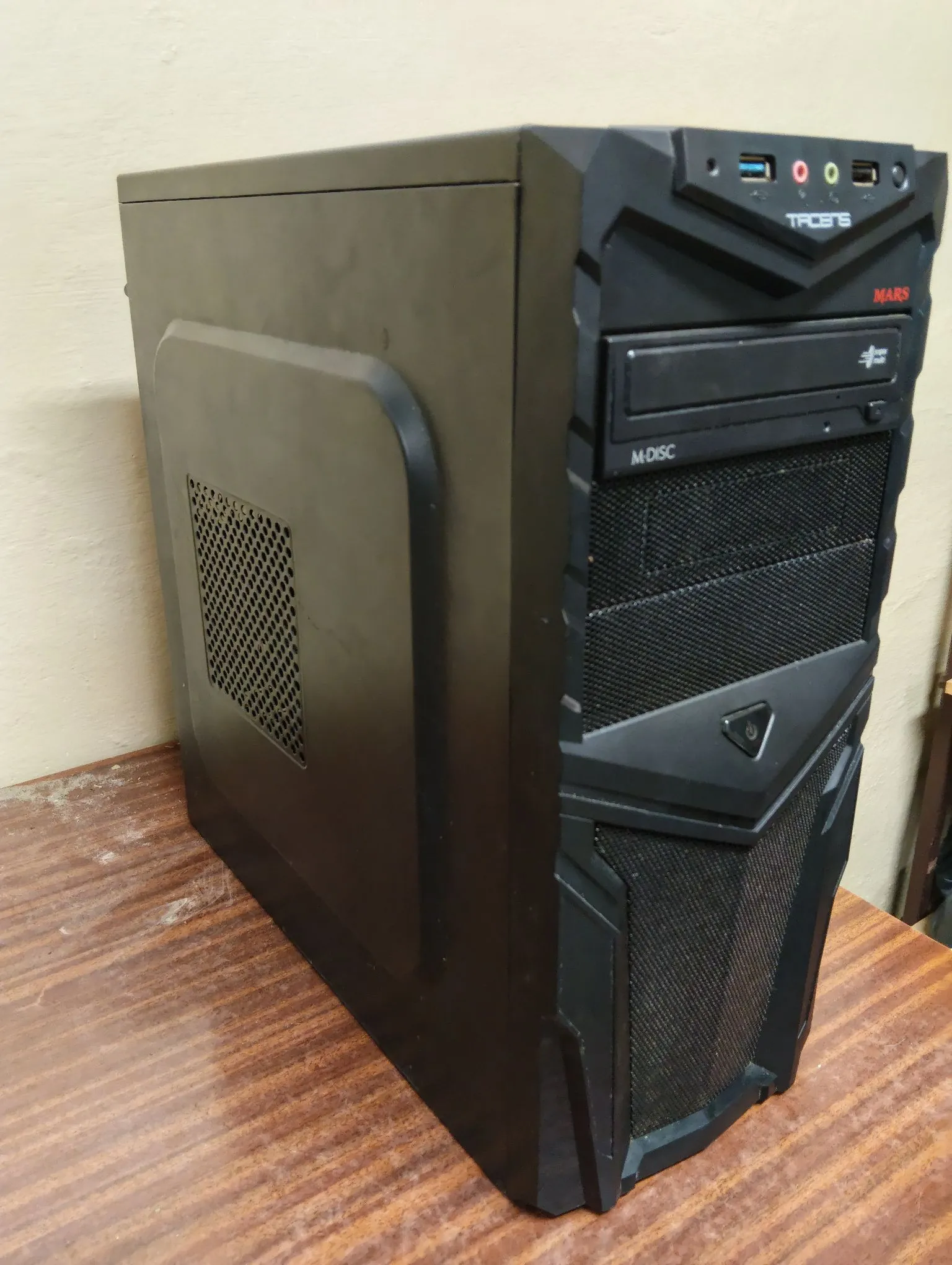 Czarna obudowa komputera stacjonarnego typu tower, ustawiona na drewnianym blacie. Na przednim panelu widoczne są porty USB, gniazdo słuchawkowe, logo marki 'TRIBES', napis 'MARS' oraz napęd optyczny z oznaczeniem 'M-DISC'. Obudowa posiada charakterystyczne, kanciaste przetłoczenia i kratki wentylacyjne.