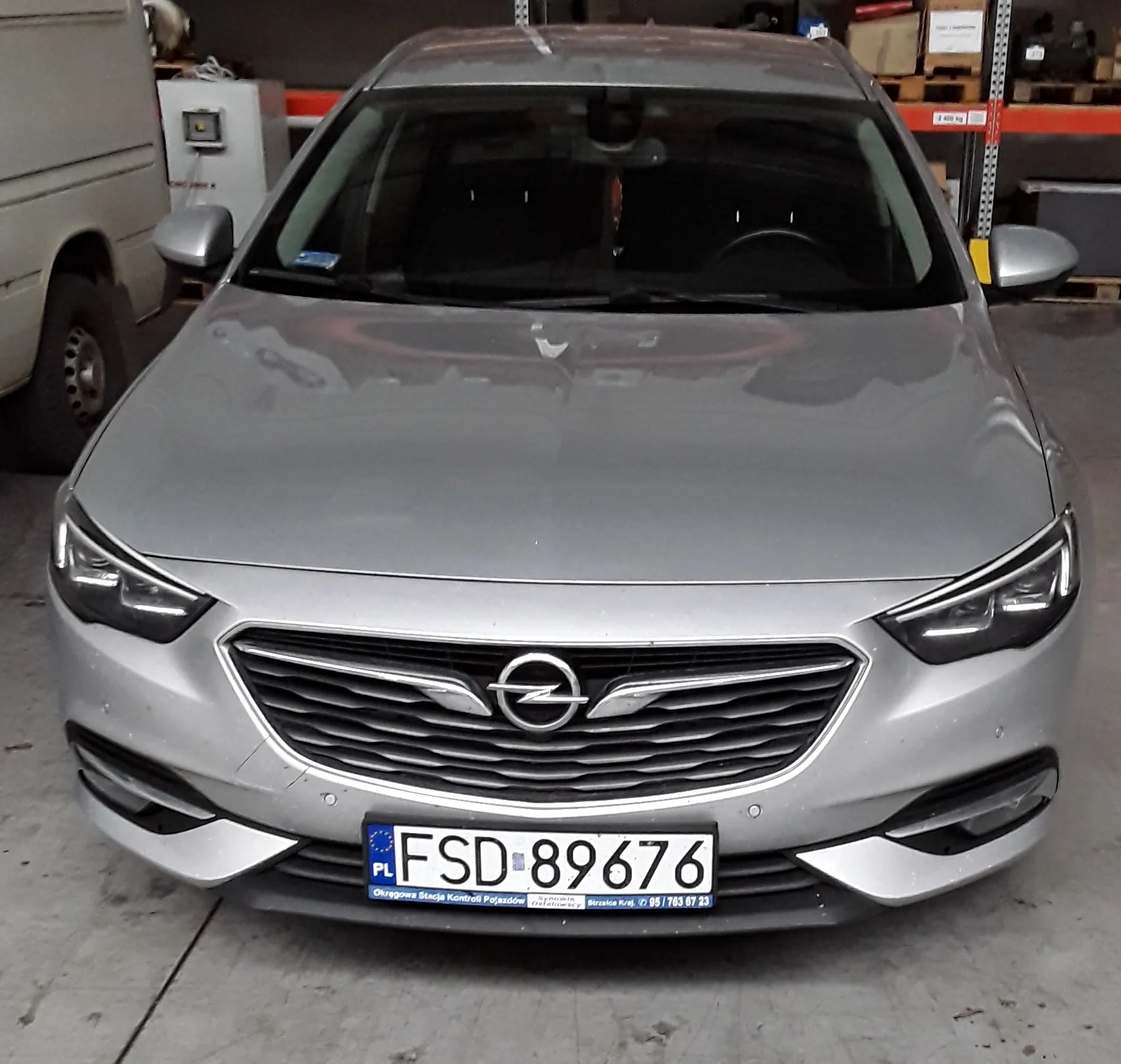 Srebrny samochód marki Opel zaparkowany w garażu lub warsztacie. Widok z przodu. Pojazd posiada polską tablicę rejestracyjną o numerze FSD 89676. Poniżej tablicy znajduje się niebieski pasek z napisem: 'Okręgowa Stacja Kontroli Pojazdów'. W tle widoczny jest biały furgon oraz regały magazynowe.