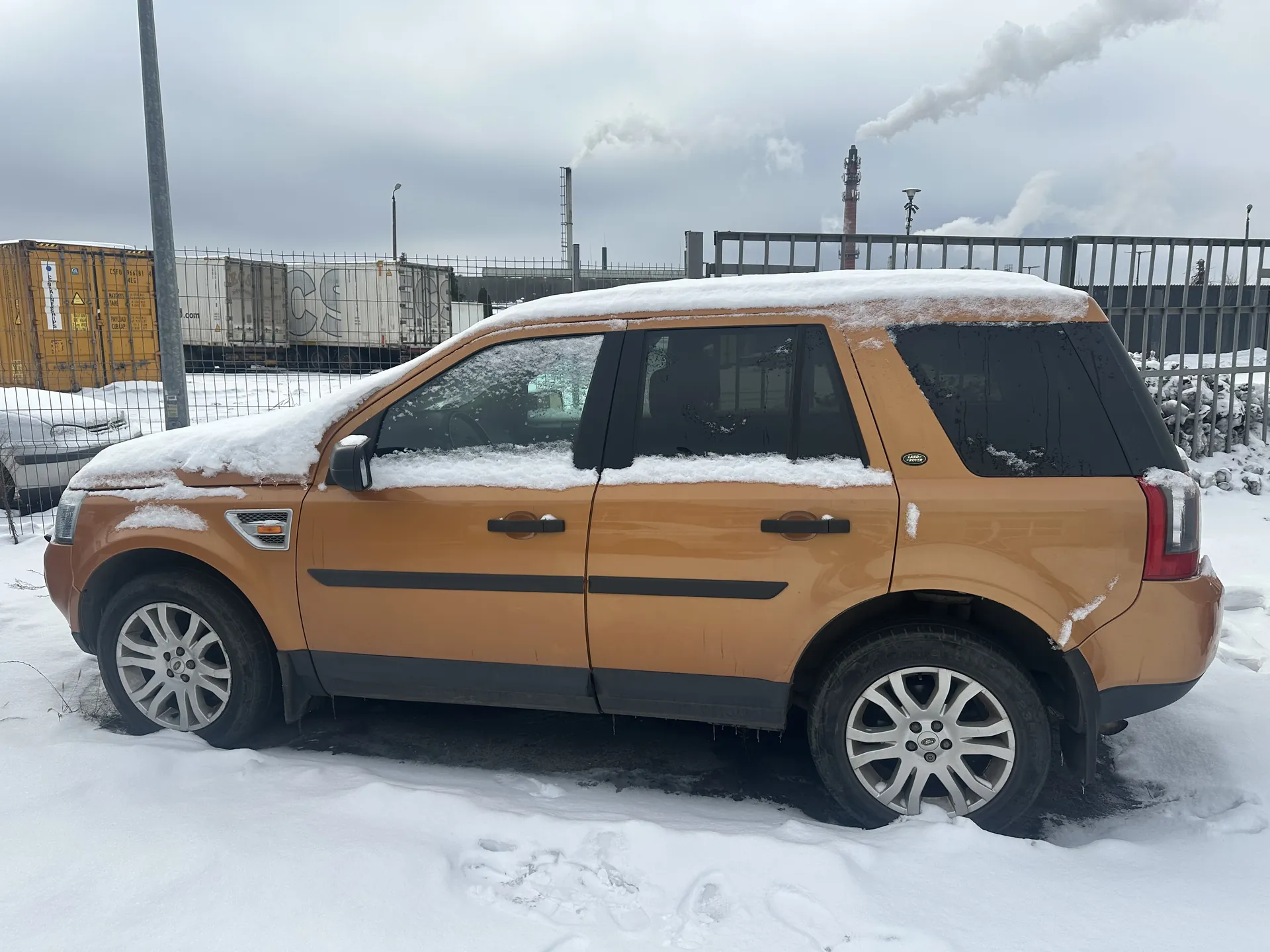 Brązowy SUV pokryty śniegiem, zaparkowany na śnieżnej powierzchni. Pojazd ma czarne słupki okienne i srebrne felgi. W tle widoczne są przemysłowe kontenery, metalowe ogrodzenie oraz kominy emitujące dym pod pochmurnym niebem. Na ziemi śnieg z widocznymi śladami stóp.