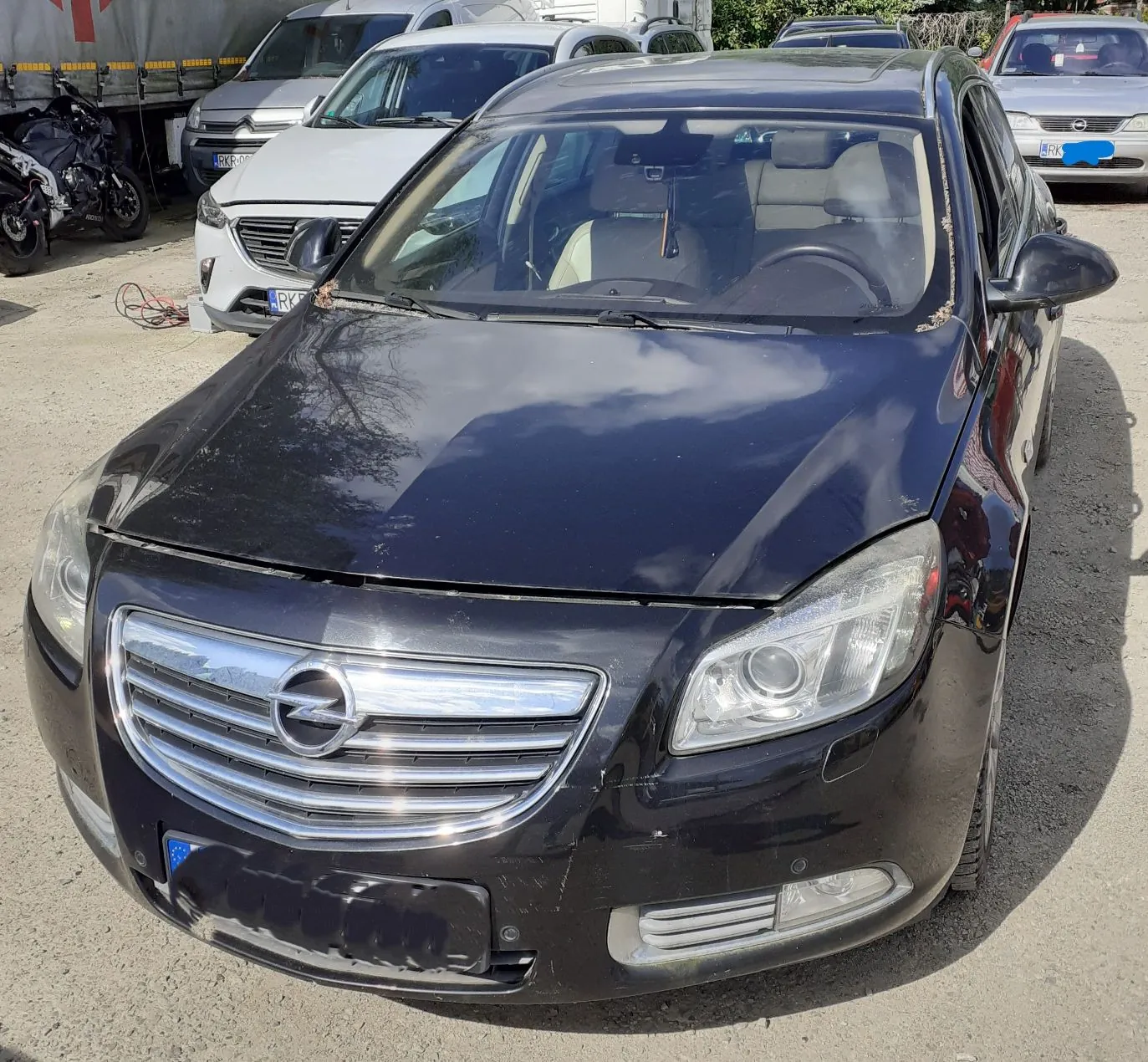 Czarny samochód marki Opel zaparkowany na placu obok innych pojazdów. Widoczny jest przód auta z charakterystycznym logo marki na chromowanej grillu. Maski samochodu są lekko uchylone, a na karoserii widać zanieczyszczenia. W tle znajdują się białe samochody oraz fragment ciężarówki.
