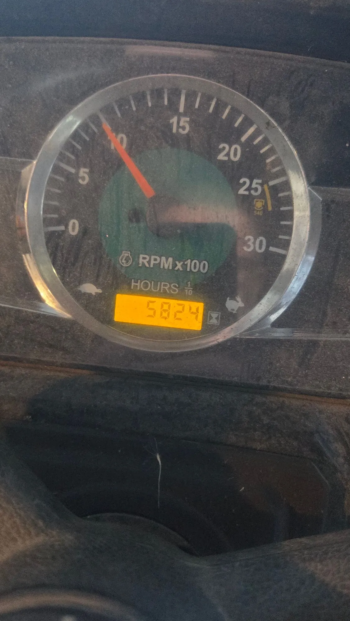 Zbliżenie na okrągły wskaźnik obrotomierza z zielonym tłem. Skala obrotów jest oznaczona jako RPM x 100, z wartościami od 0 do 30. Czerwona wskazówka znajduje się w okolicach wartości 10. Poniżej skali znajduje się napis 'HOURS 1/10' oraz cyfrowy wyświetlacz z liczbą '5824'.