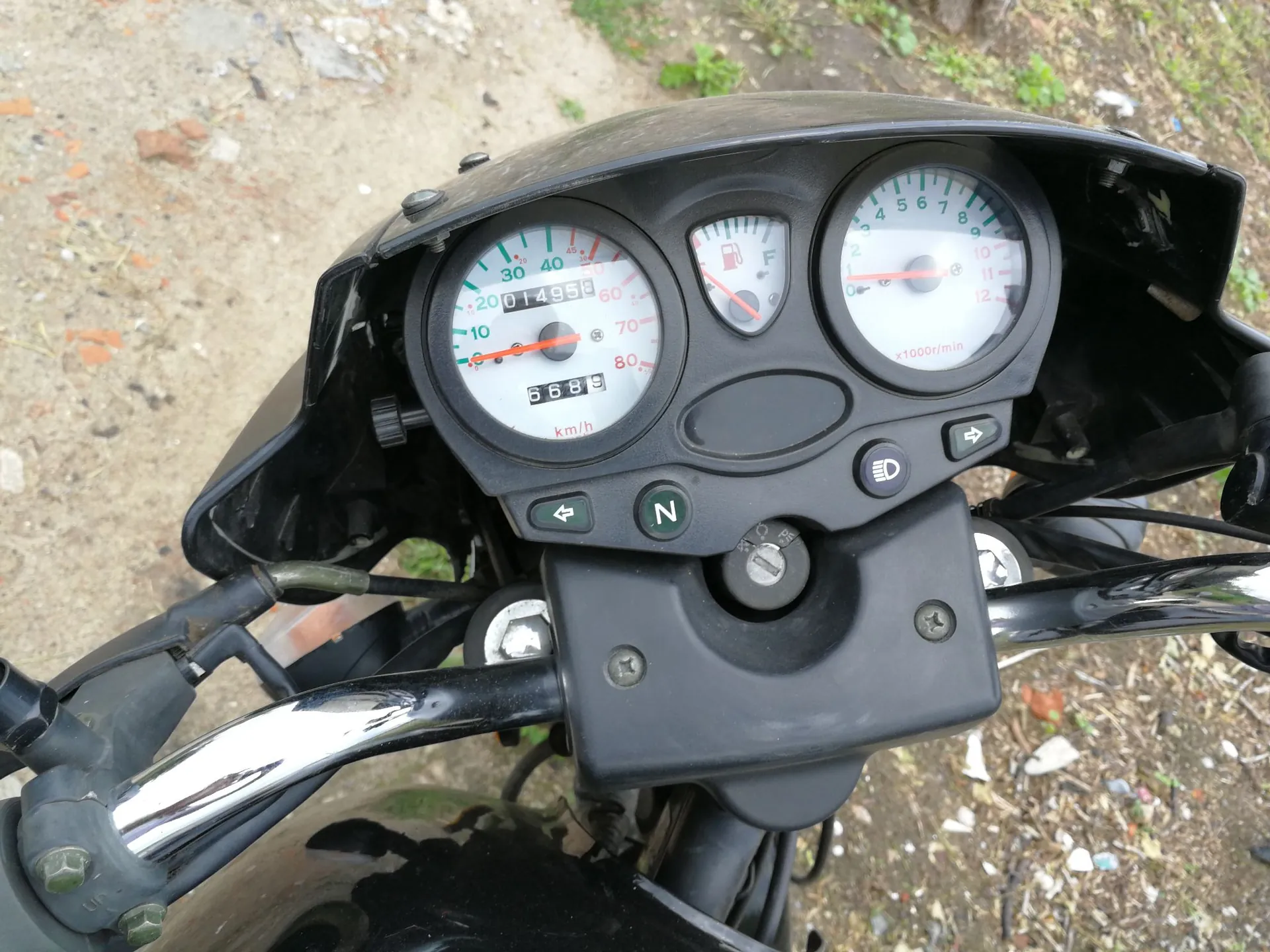 Zbliżenie na deskę rozdzielczą czarnego motocykla. Widoczne są trzy analogowe wskaźniki: po lewej prędkościomierz z oznaczeniami od 0 do 80 km/h i licznikiem przebiegu wskazującym 014959 km oraz 6689 km, w środku wskaźnik poziomu paliwa, a po prawej obrotomierz z oznaczeniami od 0 do 12 x1000r/min. Poniżej wskaźników znajdują się przyciski z oznaczeniami strzałek kierunkowych, litery N oraz ikony świateł. W dolnej części widoczna jest chromowana kierownica.