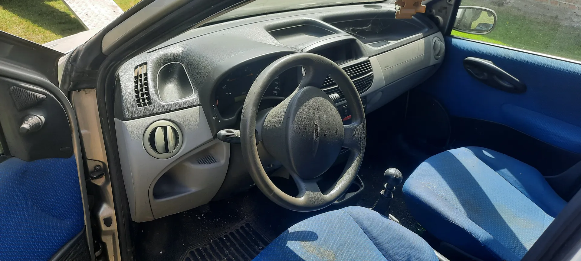 Wnętrze samochodu z niebieskimi siedzeniami, szarą deską rozdzielczą i kierownicą. Widoczne są otwarte drzwi kierowcy, deska rozdzielcza z wentylacjami, wskaźnikami prędkości, gałką zmiany biegów oraz zewnętrzny widok na trawę i chodnik. Kierownica posiada logo marki, a wnętrze wydaje się być w stanie użytkowanym z widocznymi śladami zużycia.