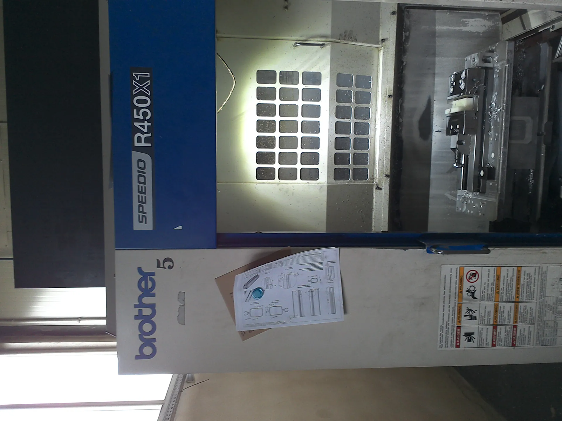 Przemysłowa maszyna marki Brother model SPEEDIO R450X1. Biało-niebieska konstrukcja z widocznym napisem 'brother' oraz cyfrą '5' po lewej stronie. Na białej części maszyny przylepiony jest dokument z rysunkami technicznymi i niebieskim klipem. Poniżej znajdują się tablice ostrzegawcze z symbolami i tekstem w kilku językach. Część robocza maszyny z siatką kwadratowych otworów, wewnątrz widoczny metalowy przedmiot z resztkami wiórów. W tle okno przepuszczające światło.