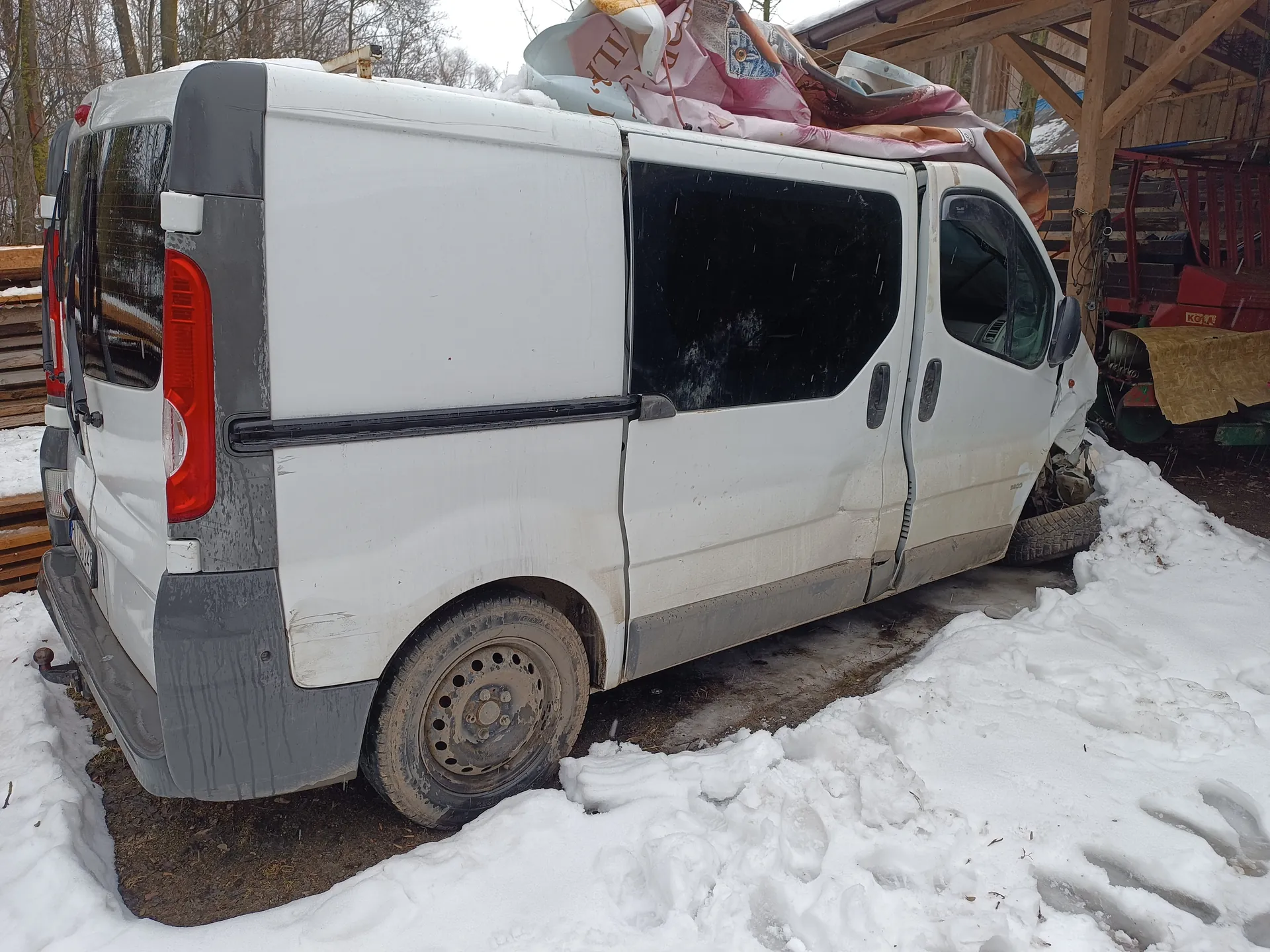Biały dostawczy van z uszkodzonym przodem po prawej stronie, zaparkowany na śniegu. Na dachu pojazdu znajdują się zakryte materiały, w tym różowa i brązowa tkanina. W tle widoczna drewniana konstrukcja garażu z czerwonymi elementami maszynowymi oraz nagie drzewa. Śnieg pokrywa ziemię, a na powierzchni widoczne są ślady stóp. Van ma zabrudzone boki i koła z brudnymi felgami.