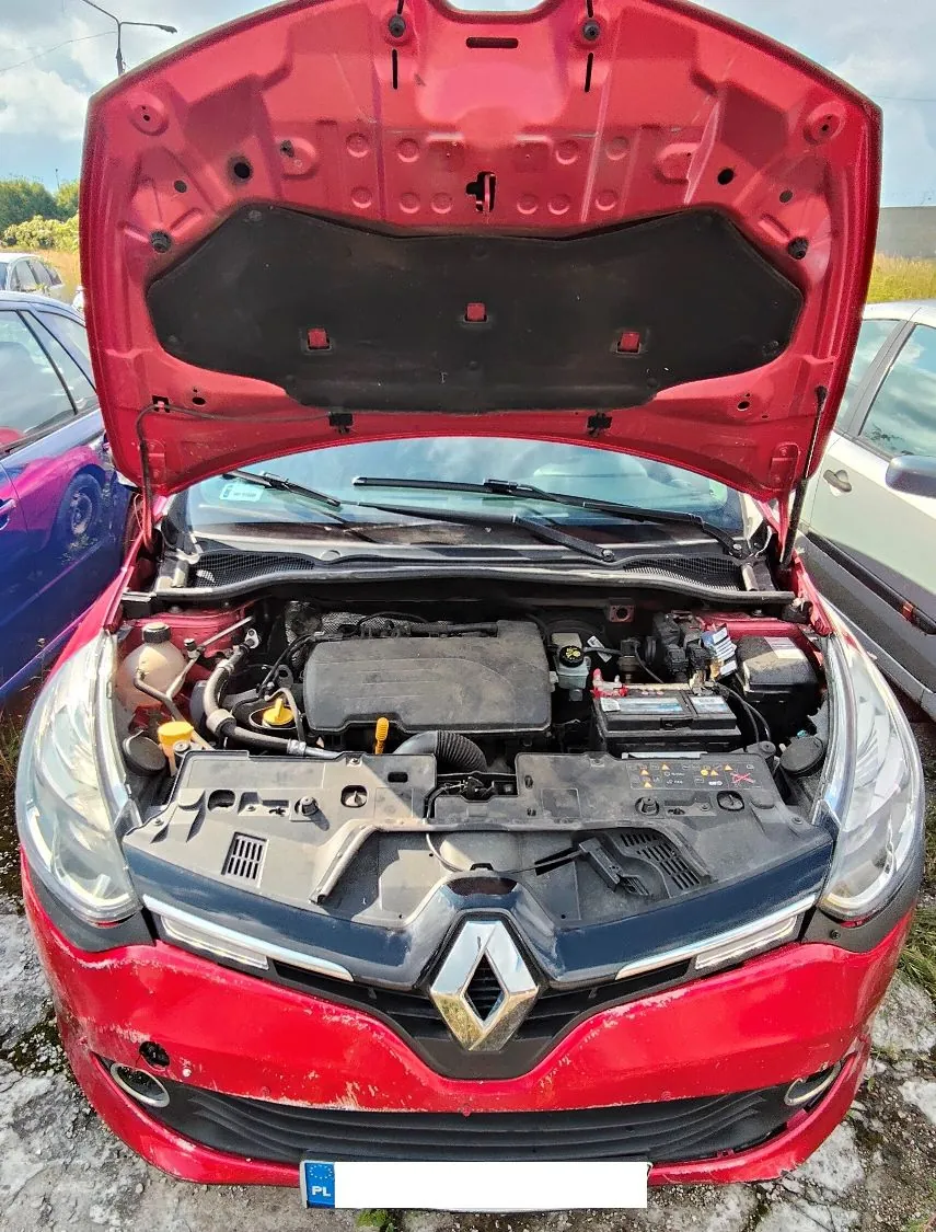 Zdjęcie przodu czerwonego samochodu marki Renault z otwartą maską, ukazującą silnik i jego komponenty. Samochód stoi na trawiastym podłożu obok niebieskiego i srebrnego pojazdu. Na przednim zderzaku widoczna jest tablica rejestracyjna z oznaczeniem PL.