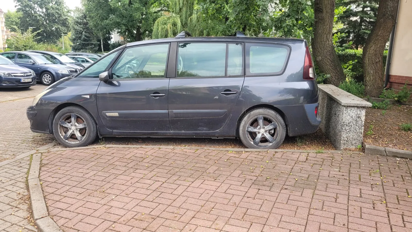 Szary samochód typu minivan zaparkowany bokiem na kostce brukowej. Pojazd znajduje się przed niskim betonowym murkiem i zielonymi drzewami. W tle widoczne są inne zaparkowane samochody.