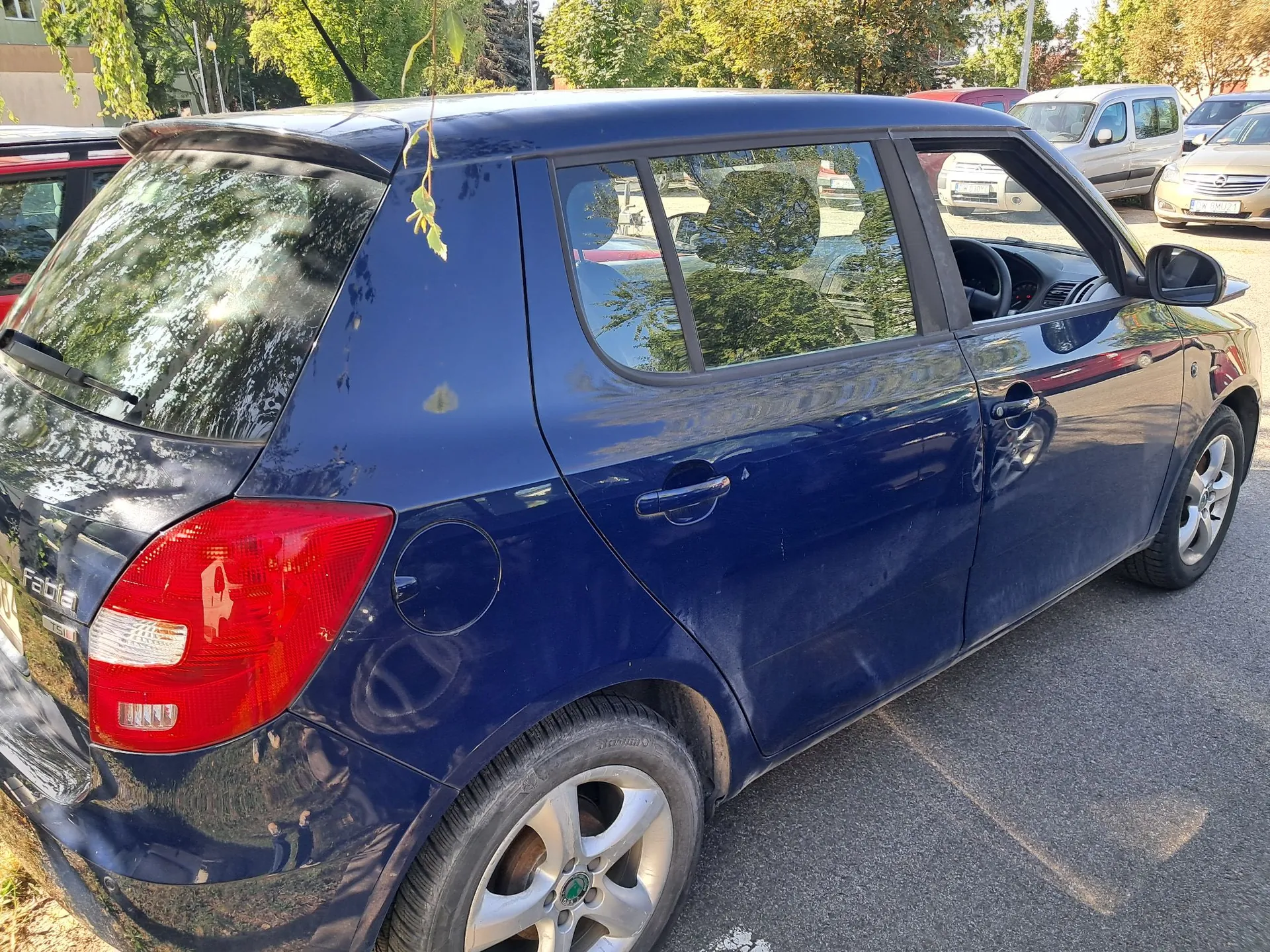 Niebieski samochód marki Skoda Fabia zaparkowany na asfaltowym placu. Widoczny jest tył i prawy bok pojazdu. Na klapie bagażnika znajduje się napis 'Fabia'. W tle widoczne są inne zaparkowane samochody oraz zielone drzewa.