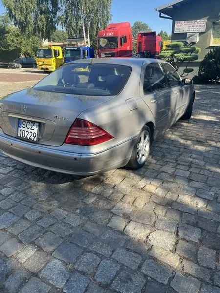 Srebrny samochód osobowy marki Mercedes-Benz zaparkowany tyłem na kostce brukowej. Na klapie bagażnika widoczna jest tablica rejestracyjna o numerze SCZ 10229 oraz napis 4MATIC. W tle znajdują się trzy ciężarówki: jedna żółta, jedna niebieska i dwie czerwone. Po prawej stronie widoczny jest fragment budynku z szyldem 'POMOC DROGOWA tel. 888 04 00 24'. W tle widać zielone drzewa i niebieskie niebo.