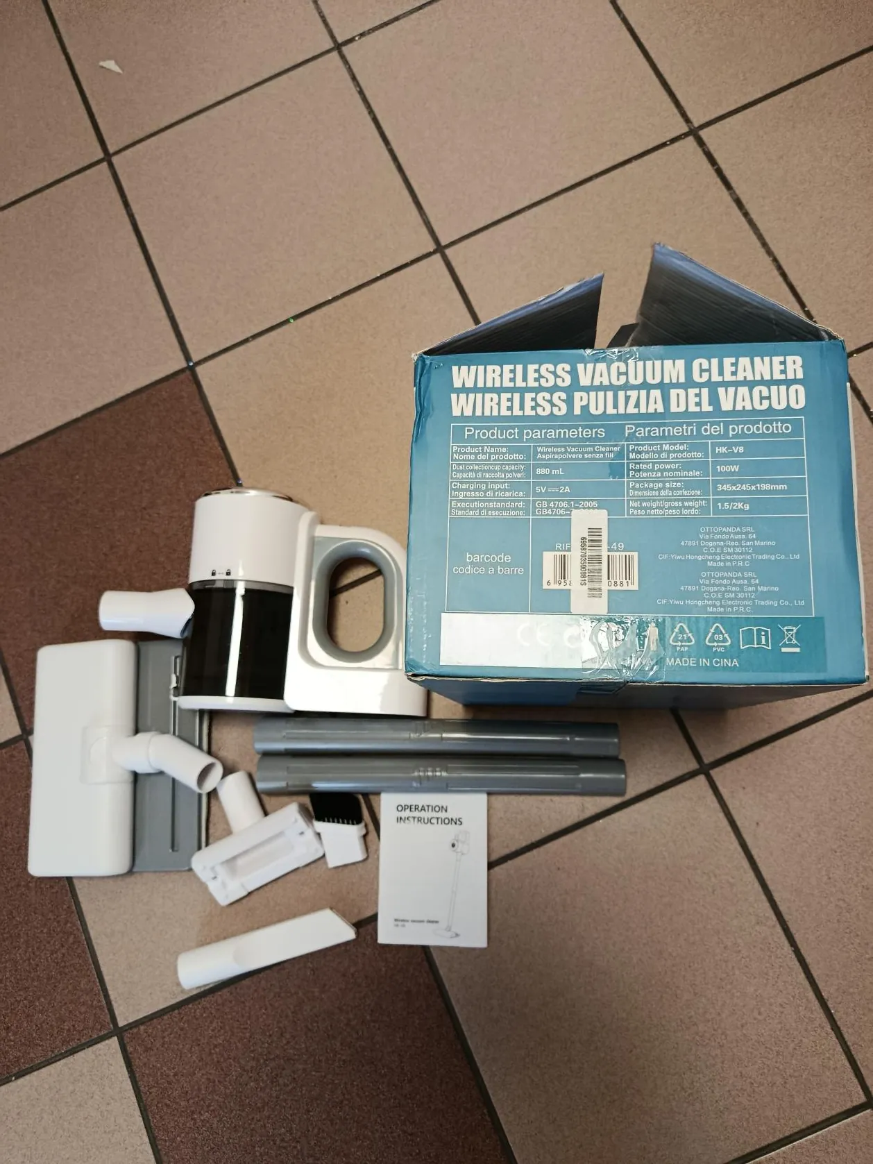 Zdjęcie przedstawia niebieskie pudełko z napisem 'WIRELESS VACUUM CLEANER' oraz 'PULIZIA DEL VUOTO' na podłodze z kafelkami w kolorach brązowym i beżowym. Obok pudełka leży biało-czarny odkurzacz ręczny, różne akcesoria (w tym szczotki, rury i podłogowa głowica), a także instrukcja obsługi z widocznym napisem 'OPERATION INSTRUCTIONS'. Na pudełku widoczne są parametry produktu, kod kreskowy oraz informacja 'MADE IN CHINA'.