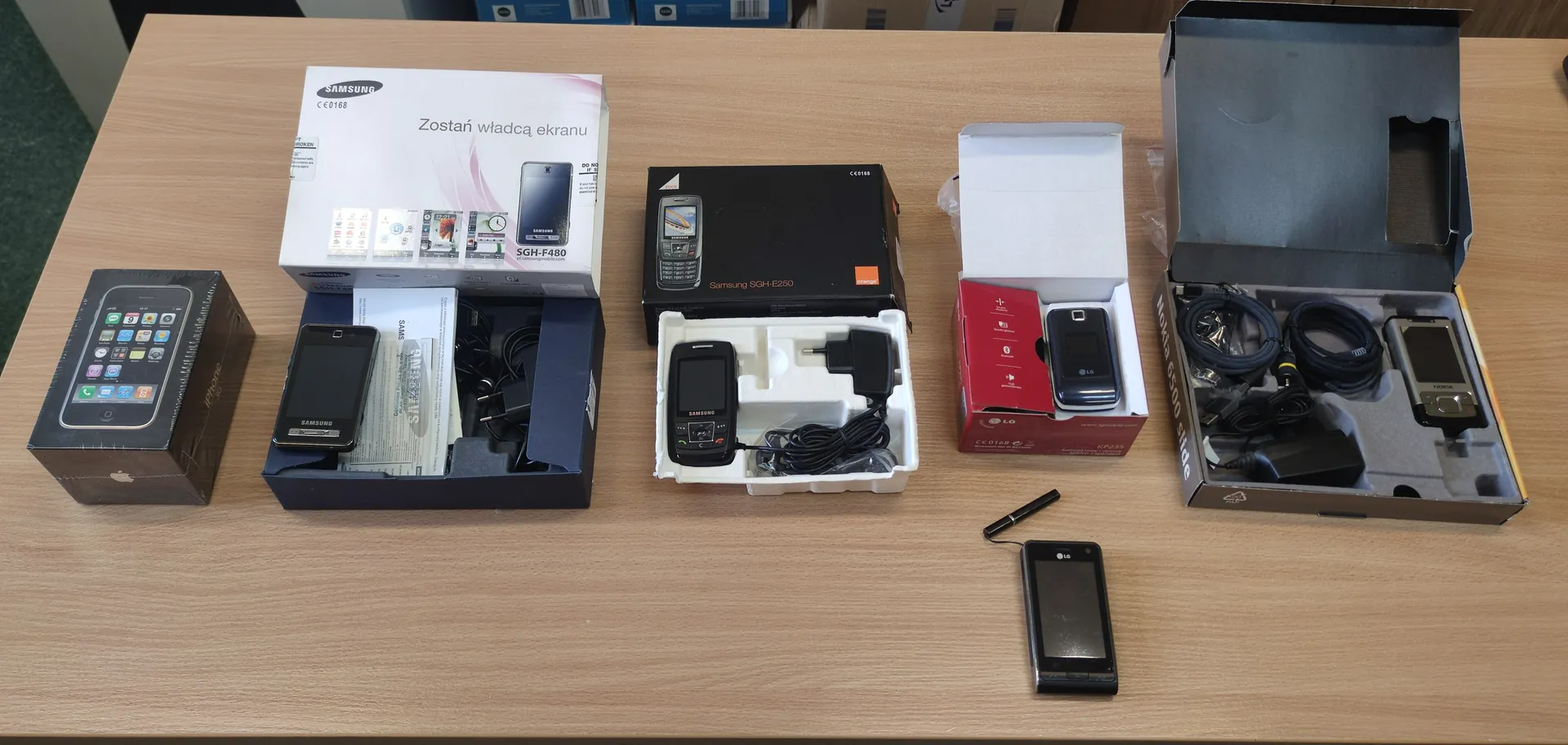 Na drewnianym stole ułożone są opakowania i urządzenia różnych telefonów komórkowych. Od lewej: czarne opakowanie z obrazem iPhone'a z logo Apple, otwarte pudełko Samsung z napisem "Zostań władcą ekranu" i modelem SGH-F480 zawierające telefon, dokumenty i akcesoria, czarne pudełko Samsung SGH-E250 z telefonem i ładowarką w białym podkładzie, czerwone opakowanie LG z telefonem wewnątrz, oraz duże otwarte pudełko z napisem "Nokia 6300 slide" zawierające telefon, kable i ładowarkę. Na stole leży również osobny telefon LG oraz długopis. W tle widoczne są niebieskie pudełka.