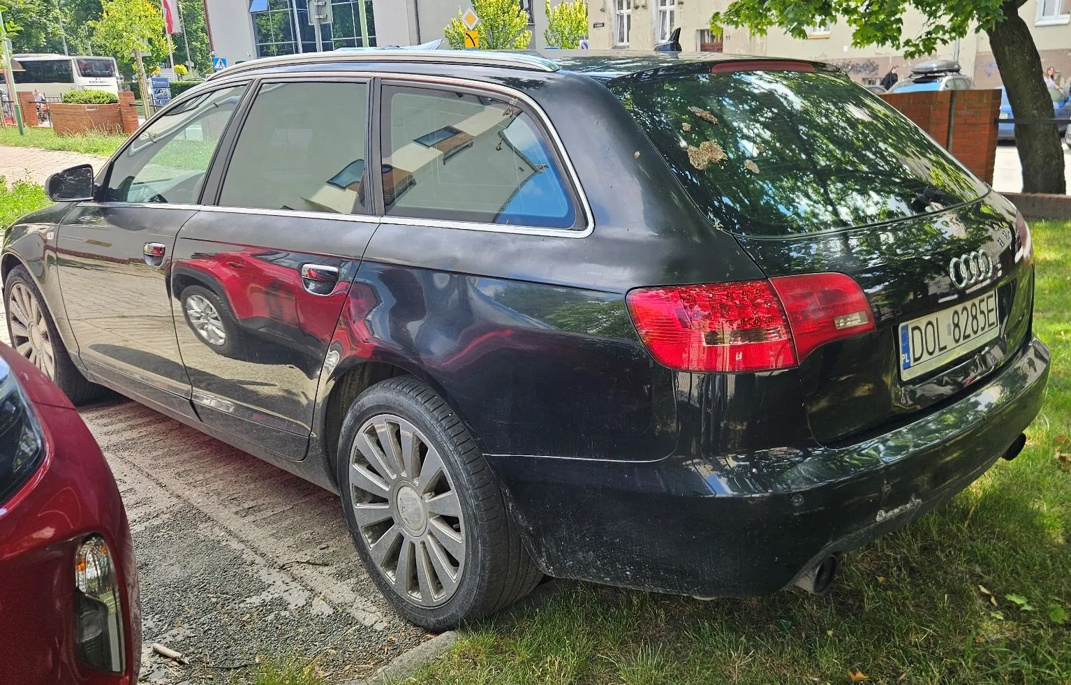 Czarny samochód marki Audi w nadwoziu kombi, zaparkowany na trawie i kostce brukowej. Widoczny jest tył i prawy bok pojazdu. Samochód ma tablicę rejestracyjną o numerze DOL 8285E. Na tylnej klapie widoczne są zacieki i uszkodzenia lakieru. W lewym dolnym rogu kadru widoczny jest fragment innego, czerwonego samochodu.