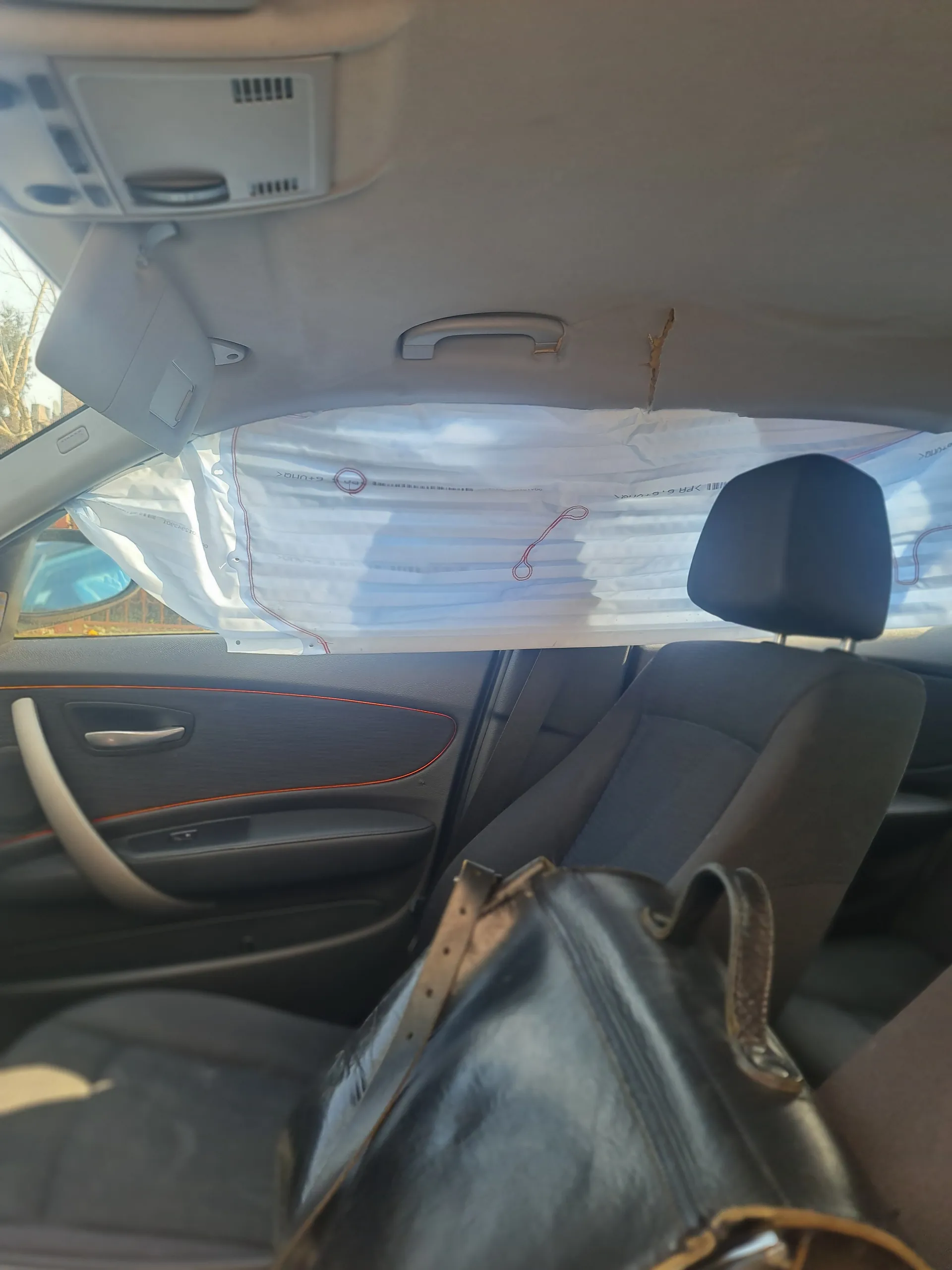 Wnętrze samochodu z widocznym lusterkiem wstecznym, czarną skórzaną torbą na siedzeniu pasażera, przezroczystą torbą na przedniej szybie z czerwonymi symbolami i częściowo widocznym tekstem, uchwytem drzwiowym, fragmentem deski rozdzielczej oraz pęknięciem na wykładzinie sufitu.
