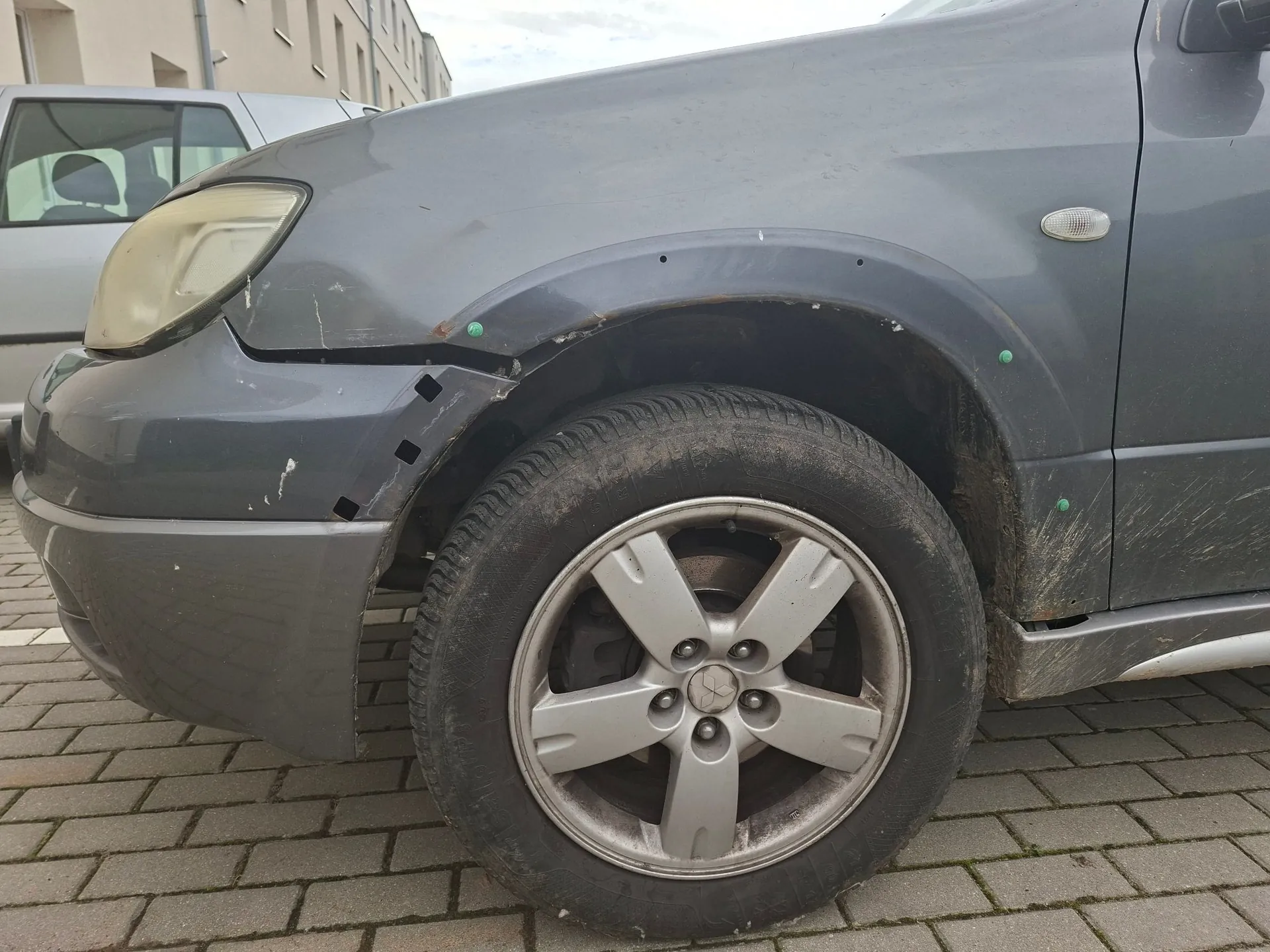 Zbliżenie na przedni prawy bok szarego samochodu z widocznymi uszkodzeniami karoserii. Przedni zderzak jest poluzowany i odstaje od nadkola, a błotnik posiada wgniecenia i zarysowania. Widoczna jest przednia lampa o zmatowiałej soczewce oraz koło z pięcioramienną srebrną felgą aluminiową. Samochód stoi na kostce brukowej.