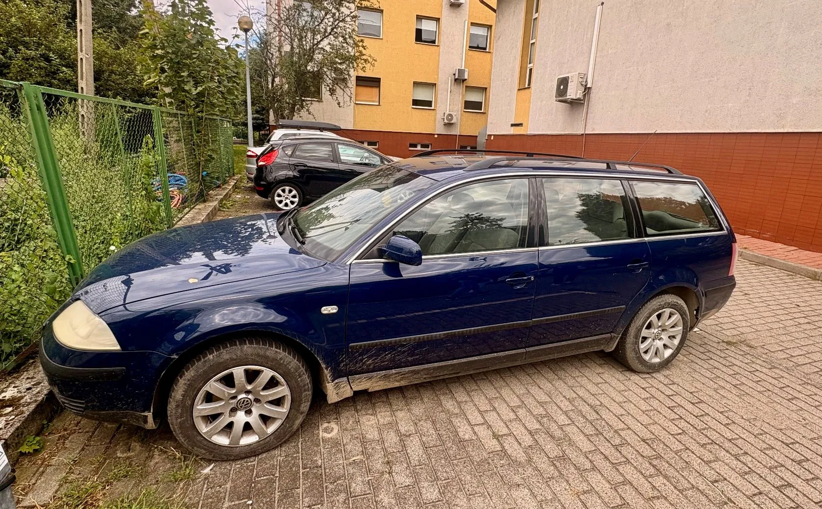 Ciemnoniebieski samochód osobowy w nadwoziu kombi zaparkowany na kostce brukowej. Auto jest zabrudzone błotem w dolnej części nadwozia. W tle widoczny jest żółty budynek mieszkalny z oknami i klimatyzatorem na ścianie oraz zielony płot z roślinnością.