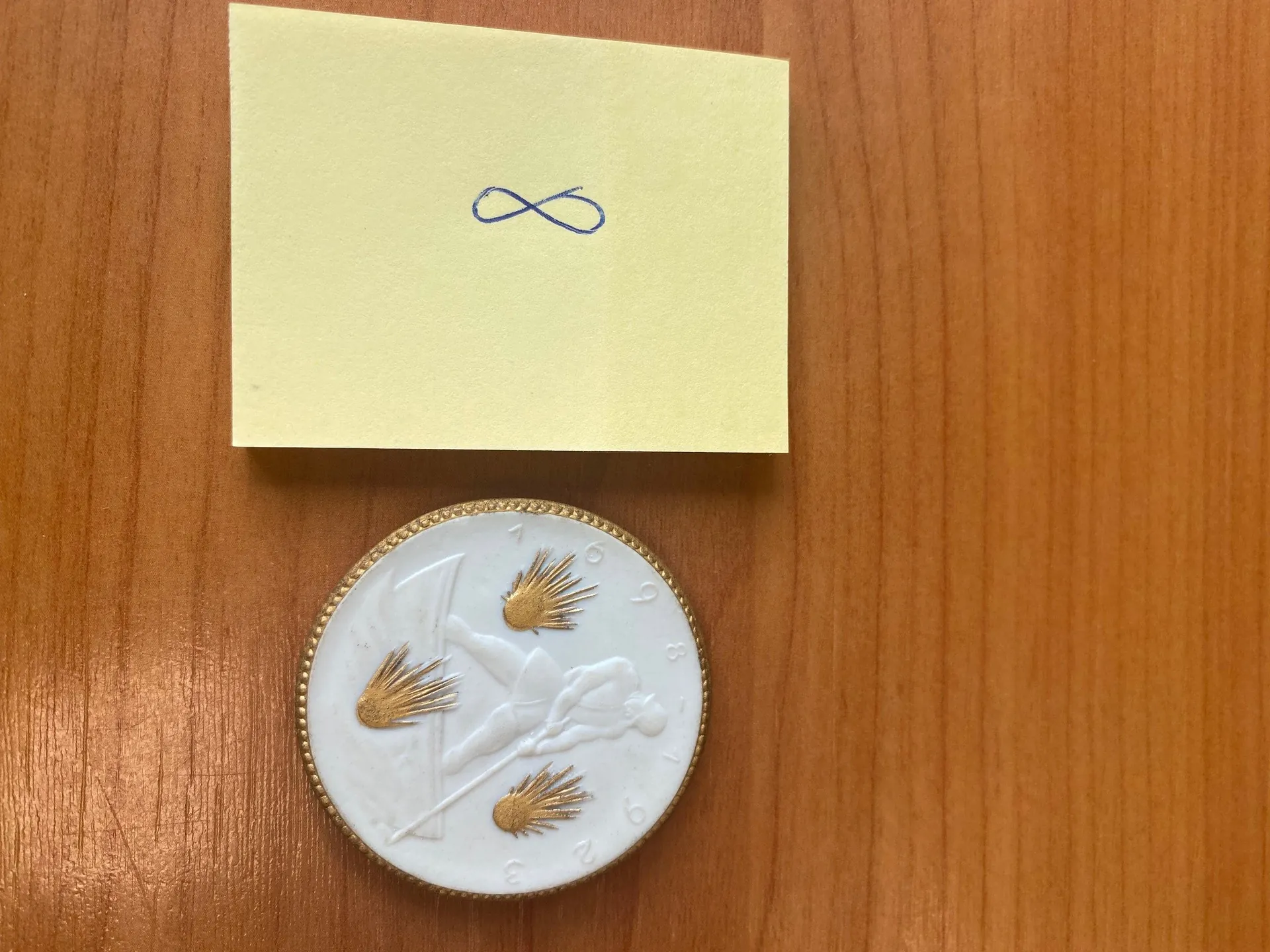 Na drewnianym tle o jasnobrązowej barwie z widocznym usłojeniem umieszczono żółty kwadratowy kawałek papieru z niebieskim symbolem nieskończoności narysowanym na nim. Poniżej papieru znajduje się okrągły przedmiot z białą powierzchnią i złotym obramowaniem, na którym widoczne są trzy złote elementy przypominające promienie słońca oraz rzeźbiona postać osoby w pozycji leżącej, trzymającej długą linię. Wokół białej części przedmiotu znajdują się rozproszone cyfry: 1, 6, 9, 3, 2, 9.