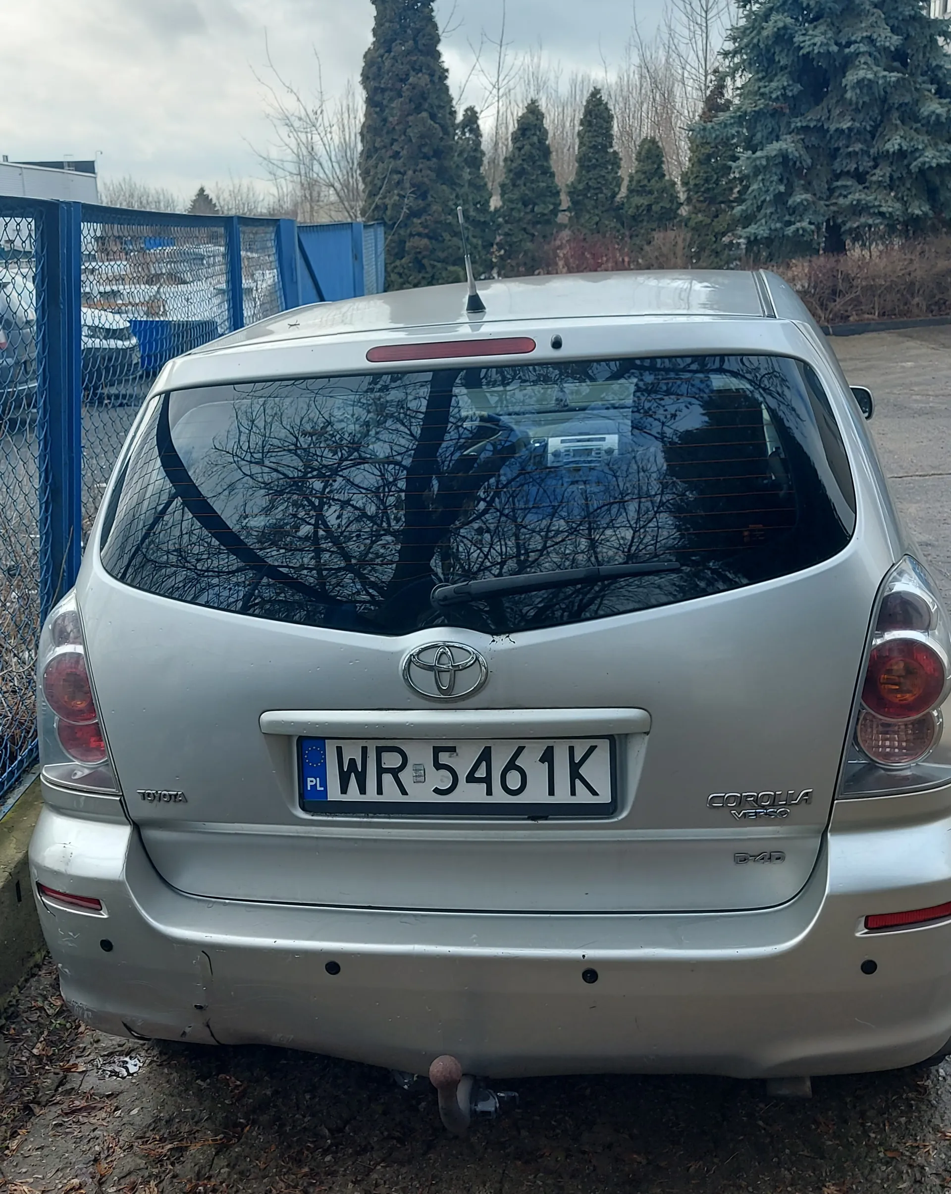 Tył srebrnego samochodu Toyota Corolla Verso z tablicą rejestracyjną WR 5461K (PL) na tle niebieskiego ogrodzenia, drzew i chmurnego nieba. Widoczne są logo Toyoty, napisy 'COROLLA VERSO' oraz 'D-4D' na bagażniku. W tle drzewa bez liści, inne pojazdy i budynki. Podwozie samochodu znajduje się na brudnej ziemi z suchymi liśćmi.
