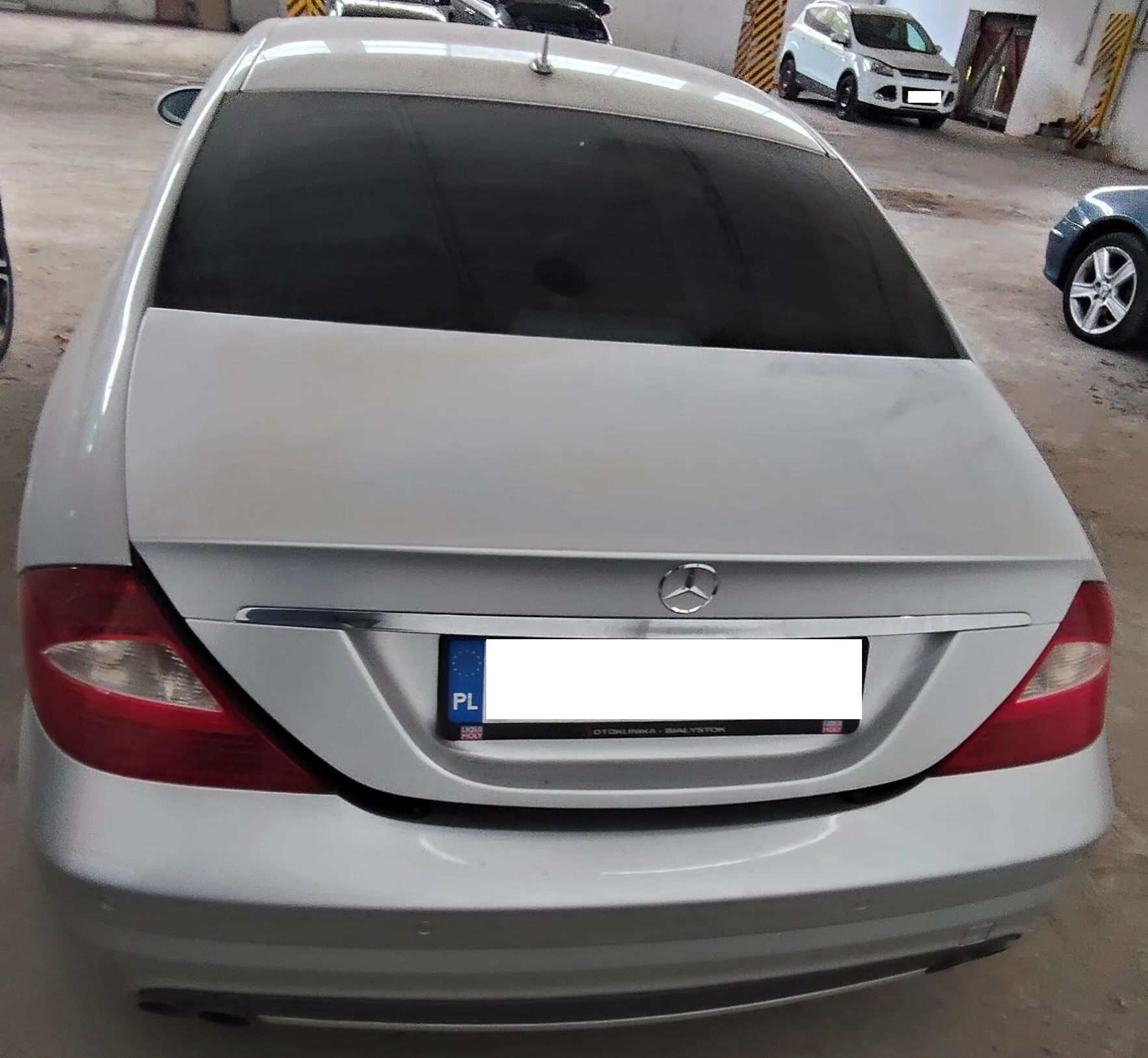Tylny widok srebrnego samochodu marki Mercedes-Benz. Widoczna jest tylna szyba, klapa bagażnika z logo marki oraz tablica rejestracyjna z oznaczeniem PL, której numer jest zakryty białym polem. Poniżej tablicy znajduje się napis 'OTOKIŃ-PIŃKA BIAŁYSTOK'. W tle widoczne są inne samochody zaparkowane na betonowym placu.