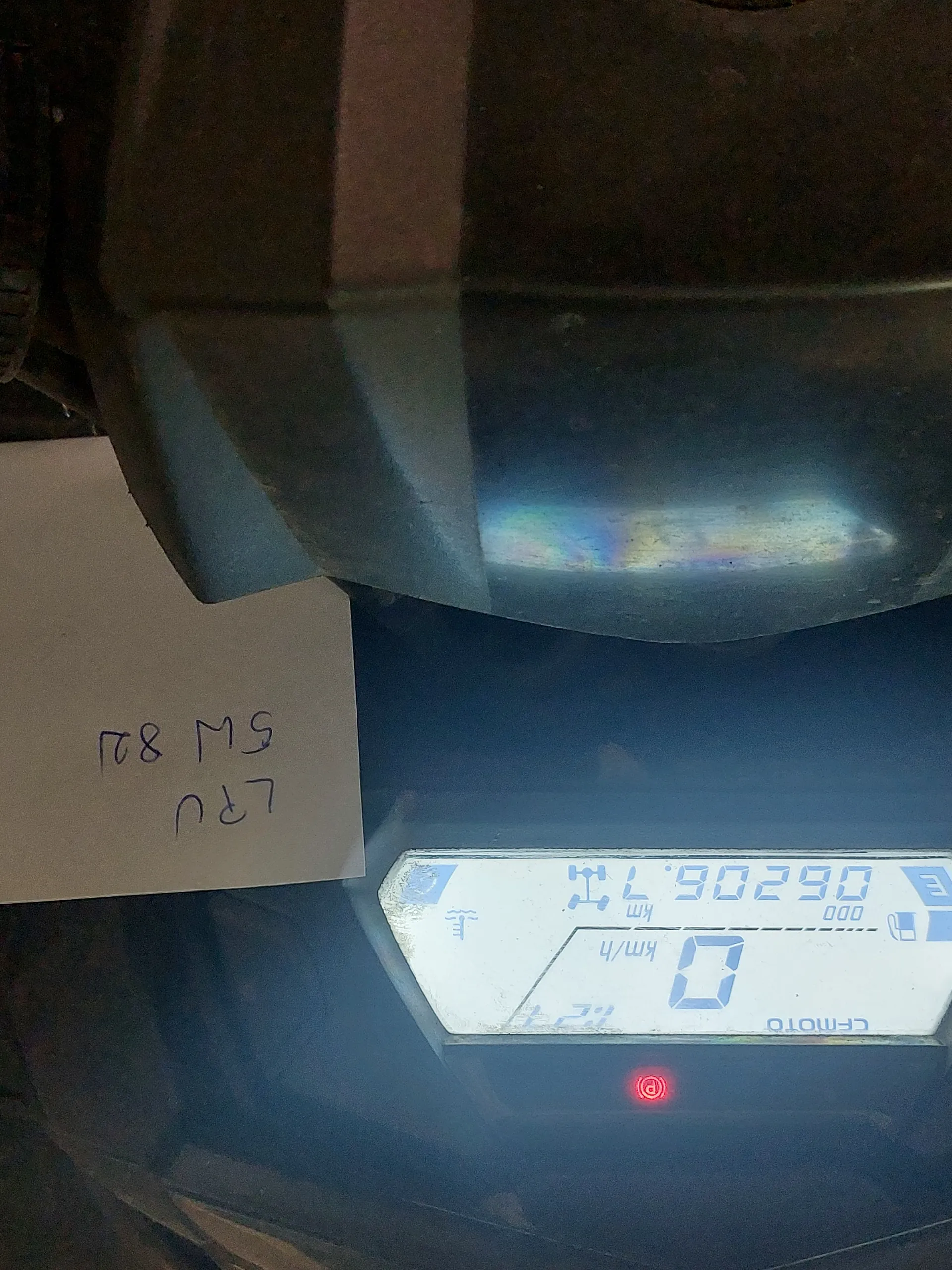 Zbliżenie na cyfrowy zegar motocykla marki CFMOTO. Wyświetlacz pokazuje prędkość 0 km/h, przebieg 06206.7 km oraz czerwone światło ostrzegawcze. Na wyświetlaczu widoczne są również symbole paliwa i termometru. Nad zegarem umieszczony jest biały kartonik z ręcznie zapisanym tekstem: LRU 5W 82J.