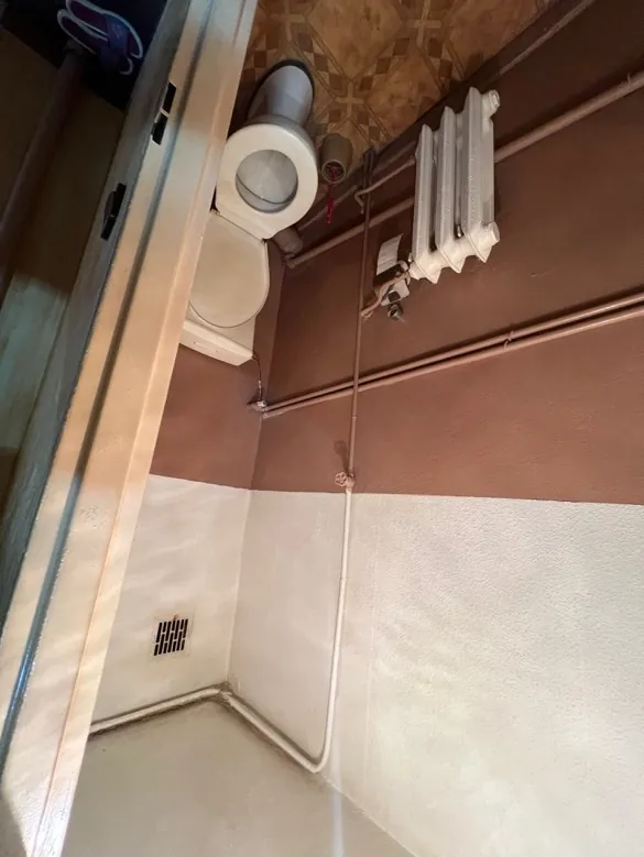 Wnętrze łazienki z białym toalet, umywalką, rurami przewodów, grzejnikiem i podłogą z wzorzystą płytką. Ściany są dwukolorowe: jasna i brązowa. Widoczna wentylacja na ścianie.