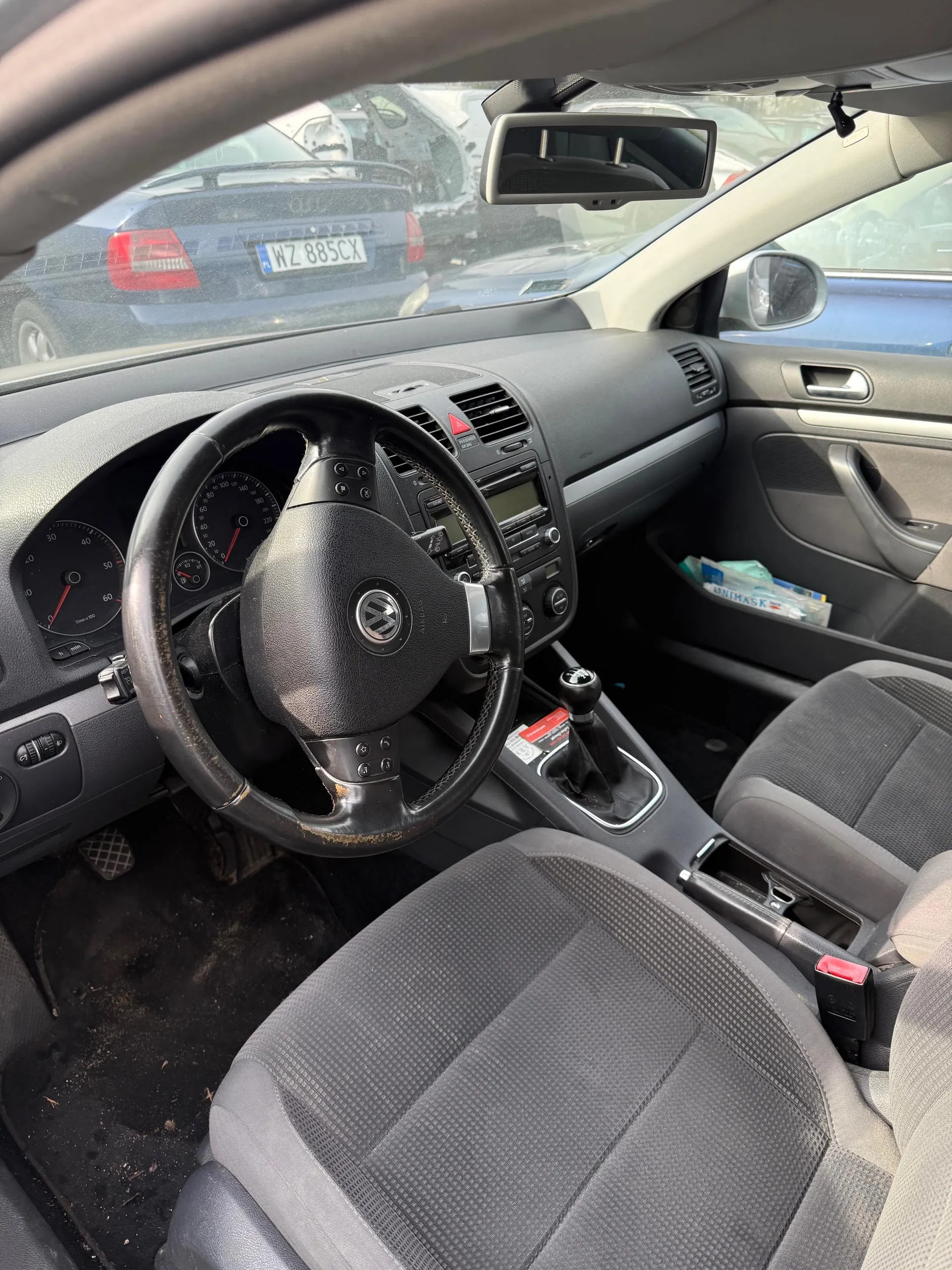 Wnętrze samochodu z widocznym kierownicą marki Volkswagen, deską rozdzielczą z licznikami, dźwignią zmiany biegów i siedzeniami w kolorze szarym. Przez przednią szybę widać inny samochód z tablicą rejestracyjną WZ 885CX. W lusterku wstecznym oraz na desce rozdzielczej widoczne są dodatkowe elementy. Podłoga w aucie jest zabrudzona.