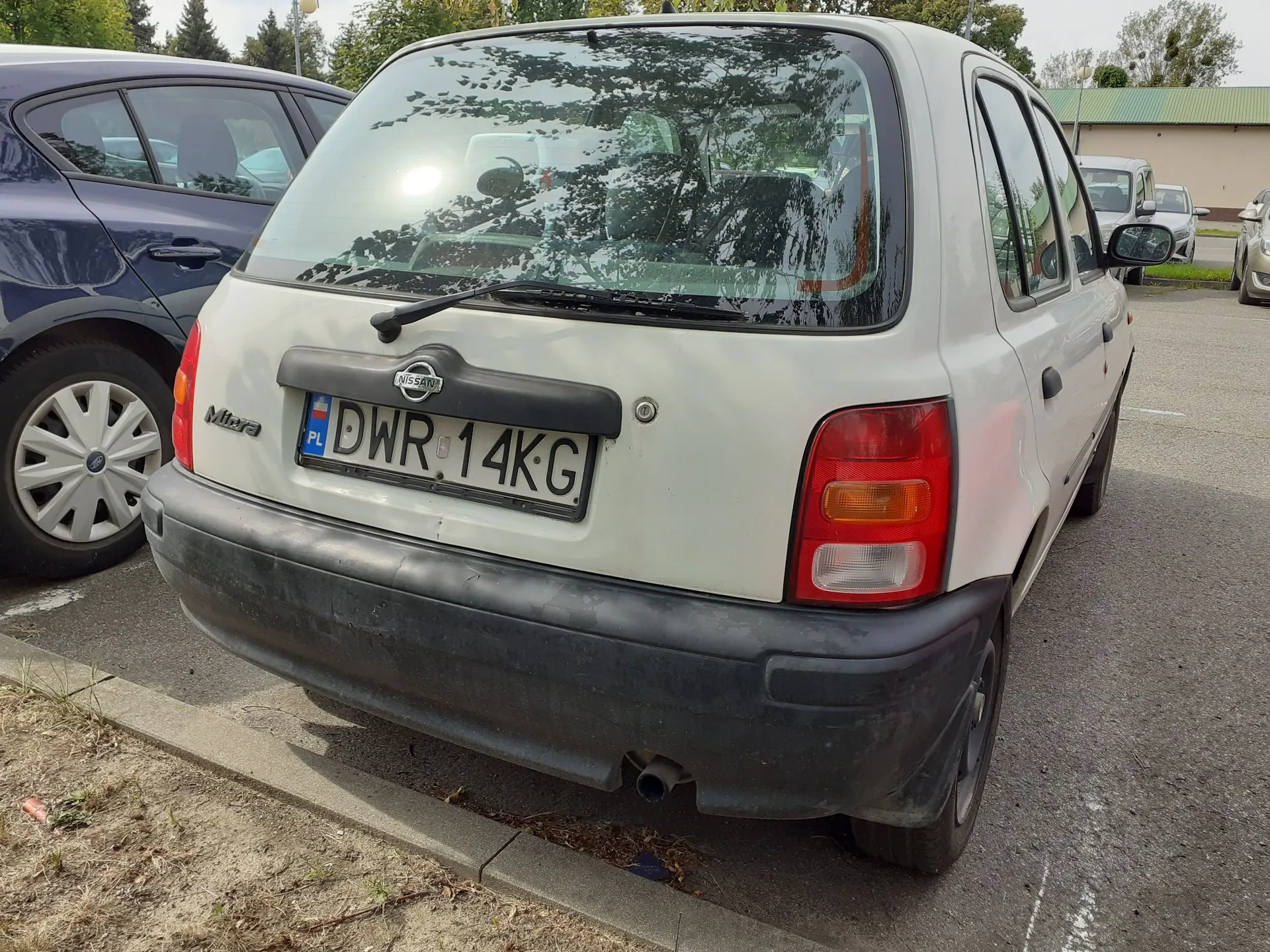 Tylny widok białego samochodu marki Nissan Micra zaparkowanego obok ciemnoniebieskiego auta. Samochód posiada polską tablicę rejestracyjną o numerze DWR 14KG. Widoczny jest czarny zderzak oraz tylne światła.
