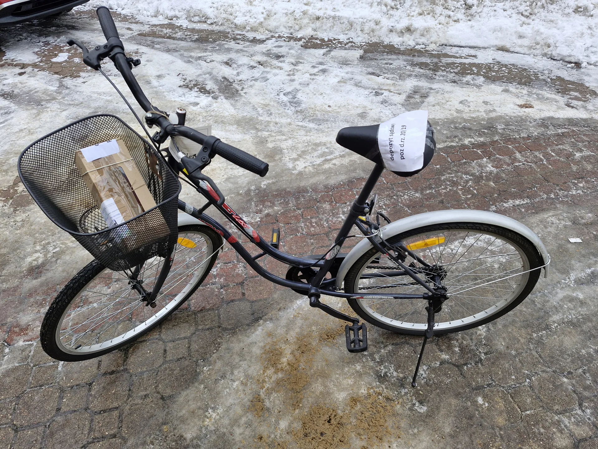 Czarny rower z koszem na przednim kole, w którym znajduje się kartonowa paczka z białymi etykietami. Na siedzeniu widoczny biały papier z napisem 'depozyt 14zł poz. d. nr 2019'. Rower stoi na brukowanej powierzchni z resztkami śniegu i lodu w tle. Widoczne są żółte odblaski na kołach oraz czarna kierownica z czarnymi uchwytami.