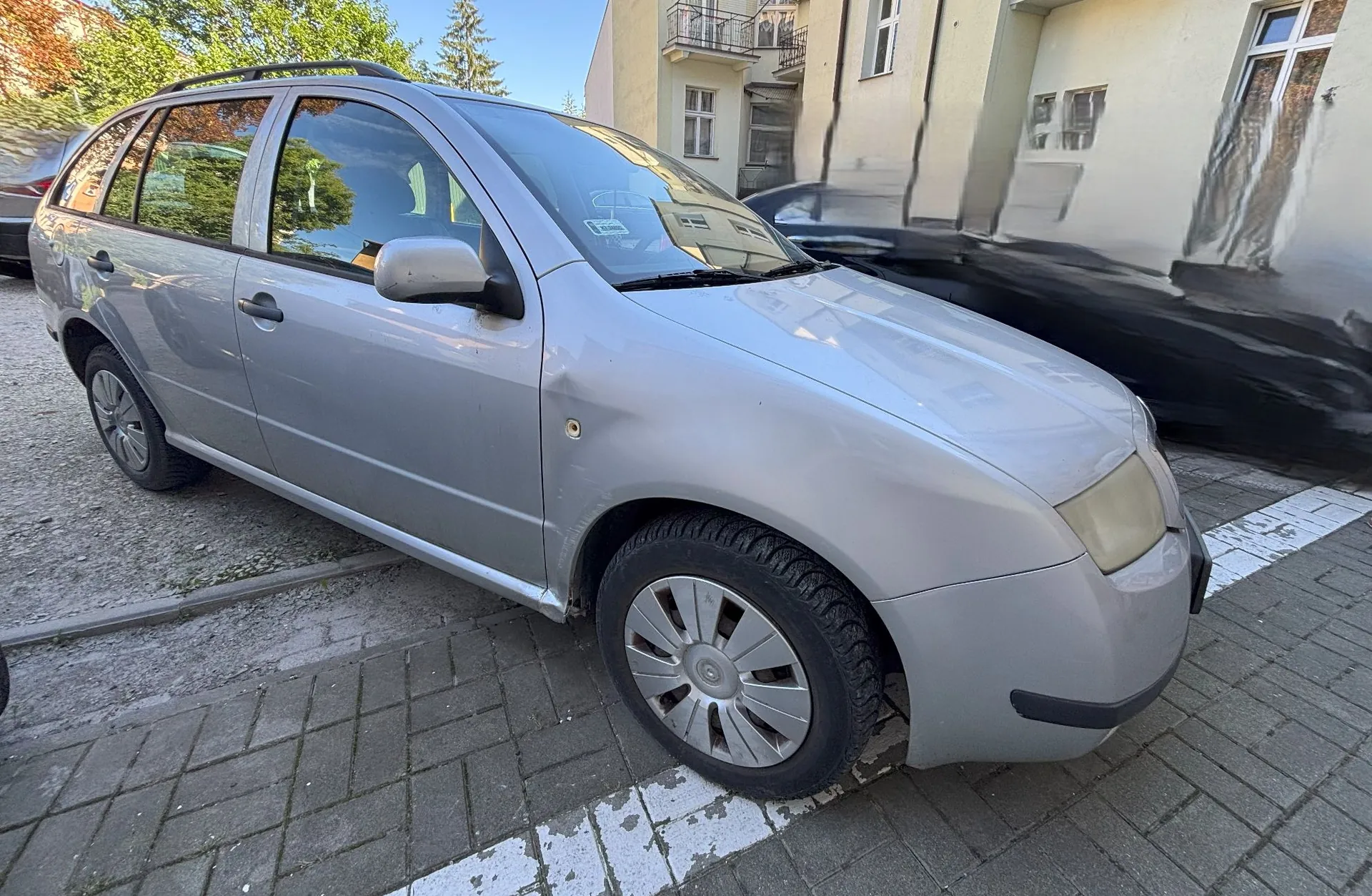 Srebrny samochód osobowy typu kombi zaparkowany na kostce brukowej. Pojazd ma widoczne wgniecenie na przednim lewym błotniku, nad kołem. W tle widać fragmenty jasnych budynków mieszkalnych oraz drzewa.