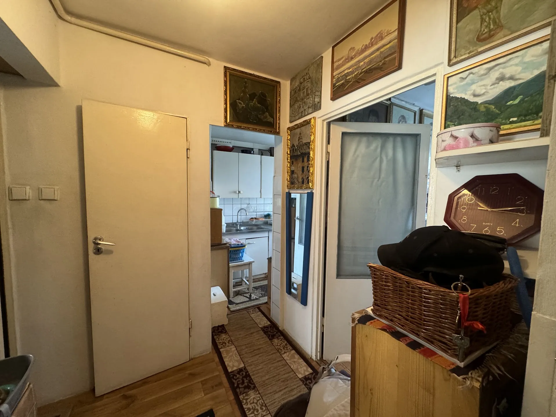Wnętrze korytarza z białymi ścianami i drewnianą podłogą. Po lewej stronie zamknięte drzwi z klamką, po prawej wiklinowy kosz na drewnianym meblu z czarną czapką i kluczami z czerwoną wstążką. Na ścianie wiszą ramkowane obrazy, lustra oraz zegar ośmiokątny z cyframi od 1 do 12. W tle widoczna kuchnia z białymi szafkami, zlewem i małym stołkiem. Na podłodze dywan z geometrycznym wzorem. Oświetlenie ciepłe, sufit biały z widoczną rurą.