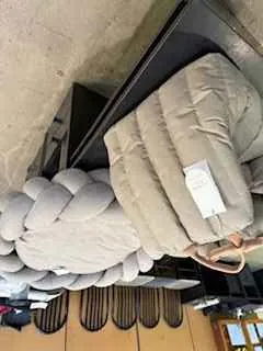 Beżowa pikowana kurtka z białą etykietą leży na czarnej powierzchni. Obok znajduje się duża, jasnoszara poduszka w stylu plecionym. W tle widoczne są drewniane krzesła z zakrzywionymi oparciami oraz betonowa ściana. Kurtka posiada brązowy pasek, prawdopodobnie część torebki.