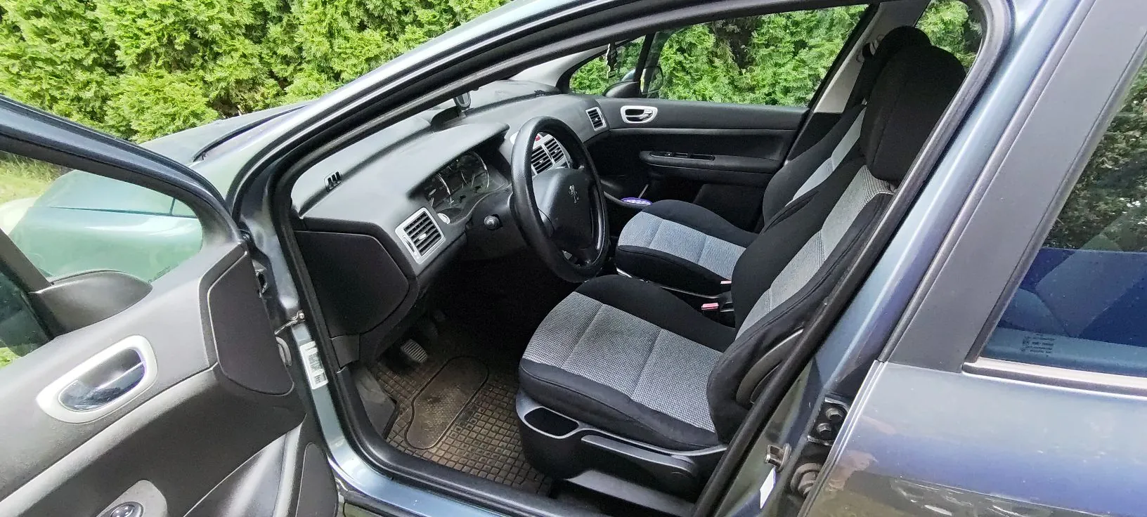 Widok z otwartych drzwi kierowcy do wnętrza samochodu. Widoczny jest czarny fotel kierowcy i pasażera z szarymi wstawkami, kierownica z logo marki Peugeot, deska rozdzielcza oraz podłoga z gumową wycieraczką. Samochód ma ciemnoszary kolor lakieru.
