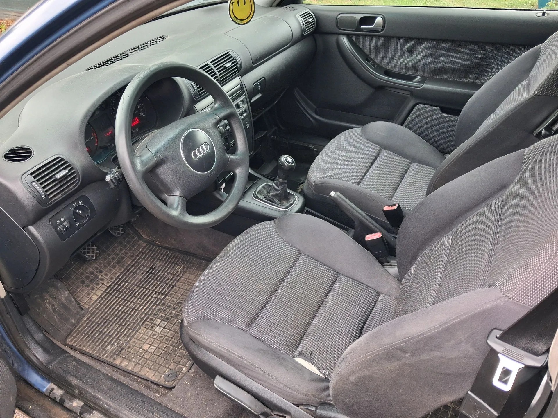 Wnętrze samochodu marki Audi widziane z perspektywy otwartych drzwi kierowcy. Widoczna jest czarna tapicerka materiałowa na przednich fotelach, czarna kierownica z logo Audi, deska rozdzielcza z zegarami i nawiewami oraz gumowa mata na podłodze. W górnej części obrazu, przy lusterku, wisi żółty brelok w kształcie uśmiechniętej buźki.