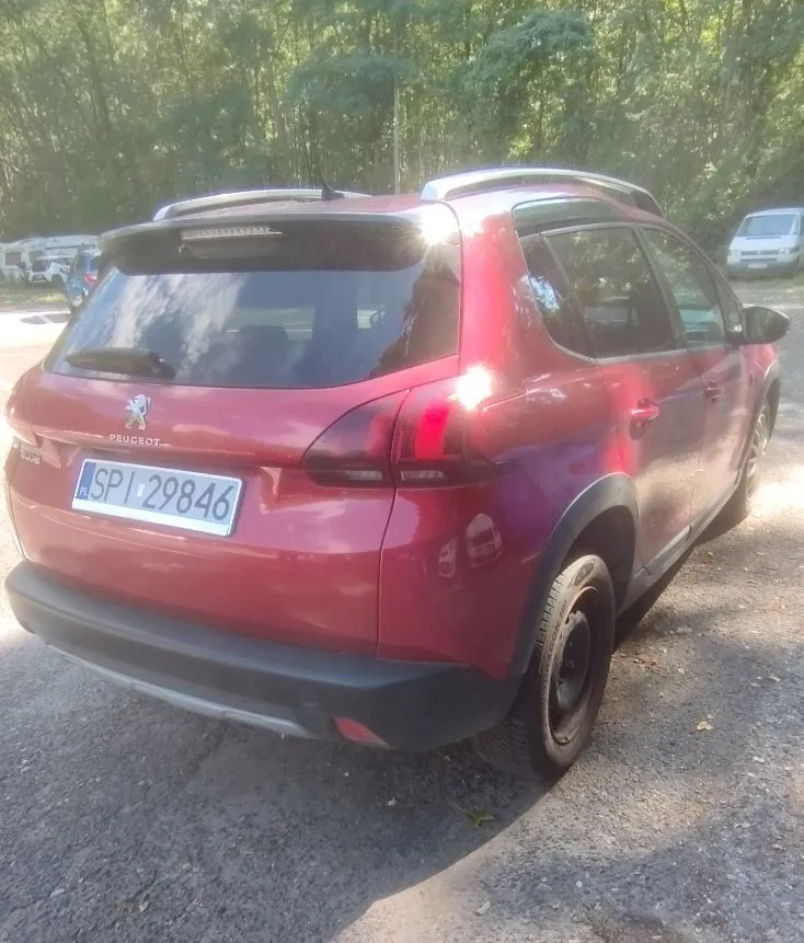 Czerwony samochód marki Peugeot w nadwoziu kombi, zaparkowany na utwardzonym placu na tle zielonych drzew. Widoczna tylna część pojazdu z tablicą rejestracyjną o numerze SPI 29846.