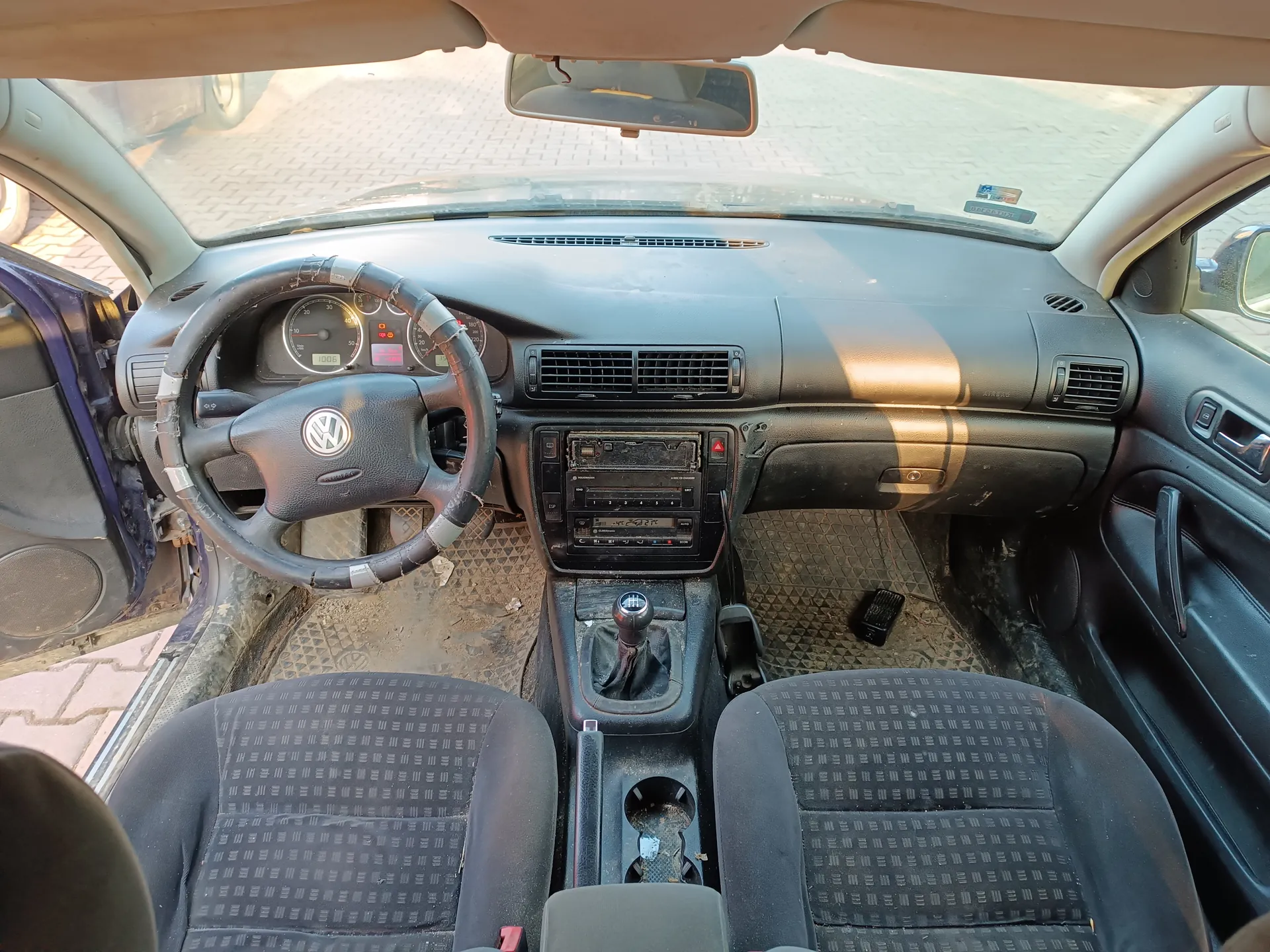 Wnętrze samochodu Volkswagen z widokiem na deskę rozdzielczą, kierownicę z logo VW, wskaźniki prędkości i obrotomierza, centralny panel z odtwarzaczem i klimatyzacją, dźwignię zmiany biegów oraz czarne siedzenia z geometrycznym wzorem. Drzwi po stronie kierowcy są otwarte, a na podłodze widoczne są brudne dywaniki. Przez przednią szybę widać brukowaną powierzchnię. Wnętrze wydaje się zabrudzone, a na desce rozdzielczej znajdują się ślady zużycia.